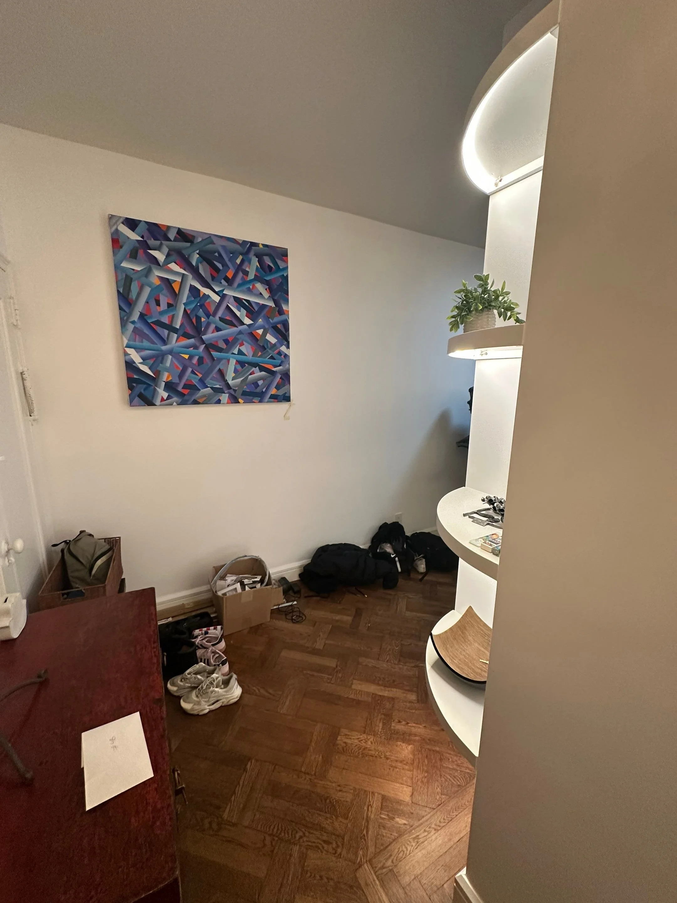 Empty room with a colorful abstract painting on the wall, hardwood floor, shoes, backpack, and some papers on a red desk, with a curved white shelf with plants and magazines.
