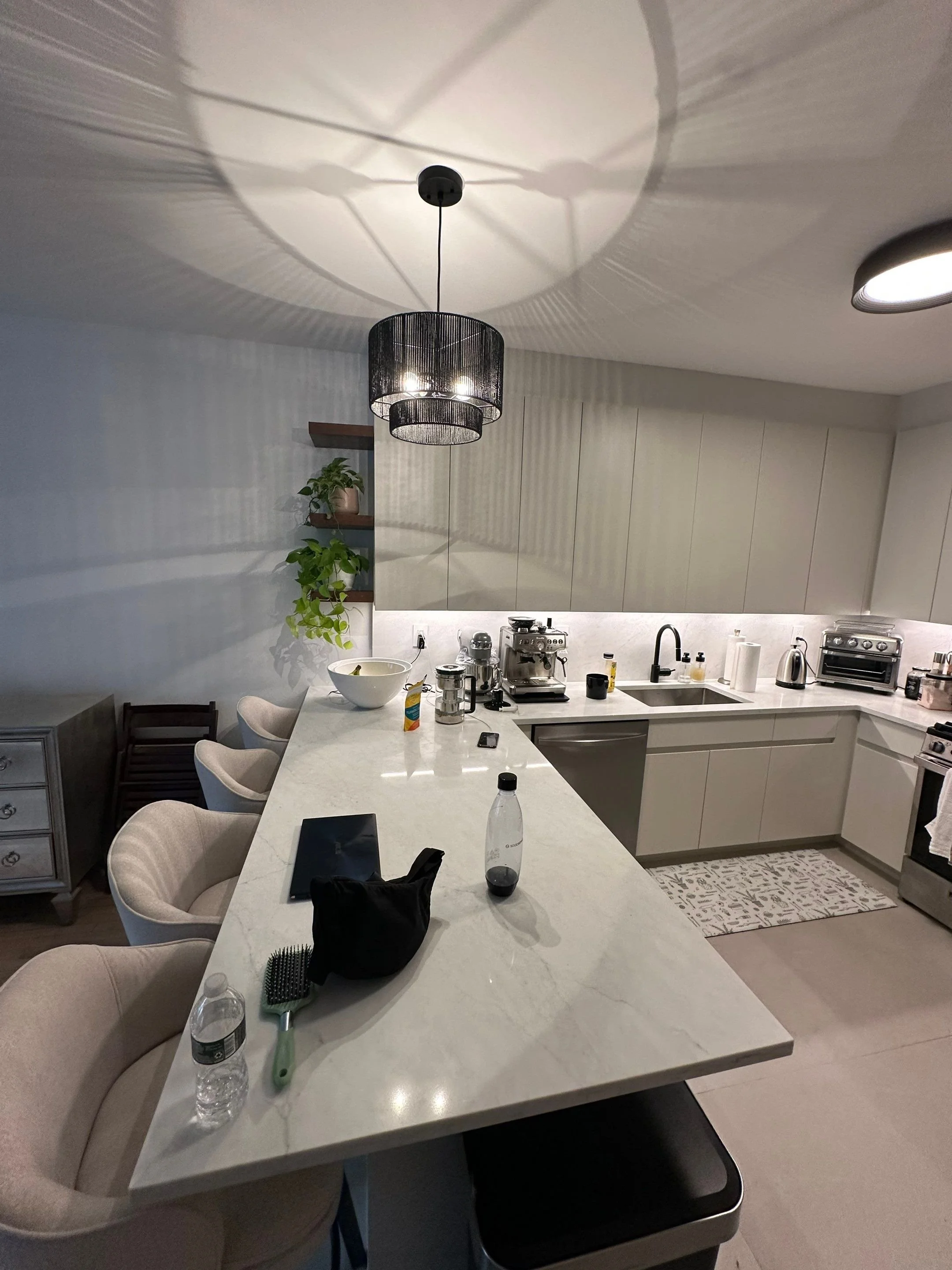 Modern kitchen with white marble island, beige chairs, black hanging pendant light, and various appliances and items on the countertop.
