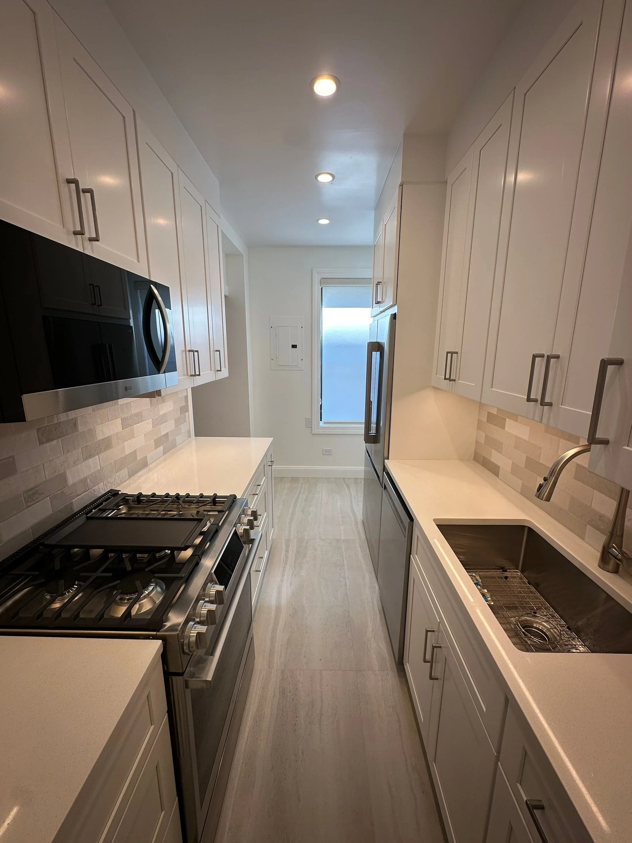 A modern, narrow kitchen with white cabinets, stainless steel appliances including a microwave and stove, a beige tile backsplash, a single sink, and a window at the end of the room.