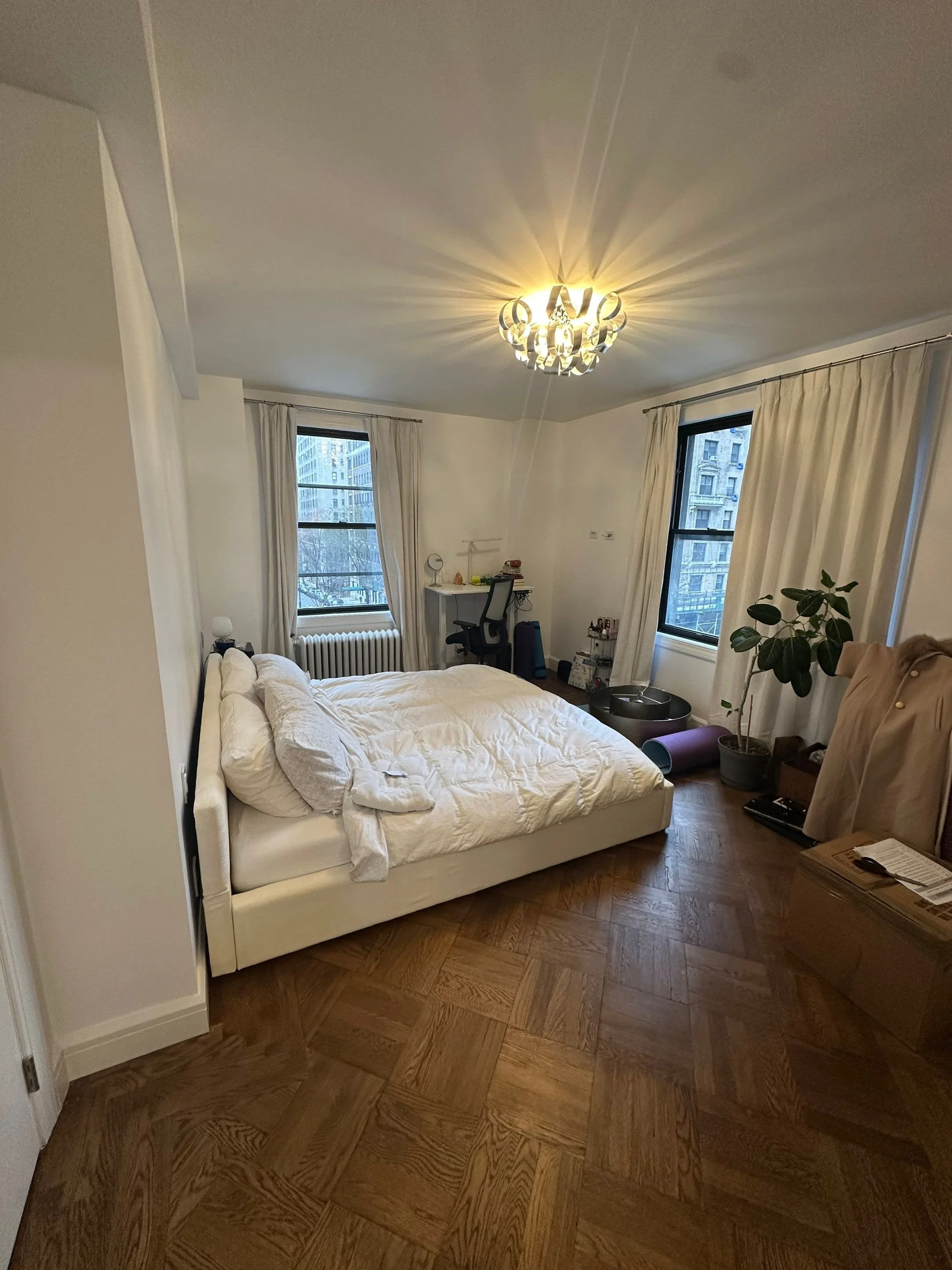 A bedroom with a white bed, two windows with curtains, a small desk with a lamp and chair, desk accessories, a cozy chair covered with a beige blanket, a potted plant, and a wooden floor. The ceiling has a decorative chandelier.