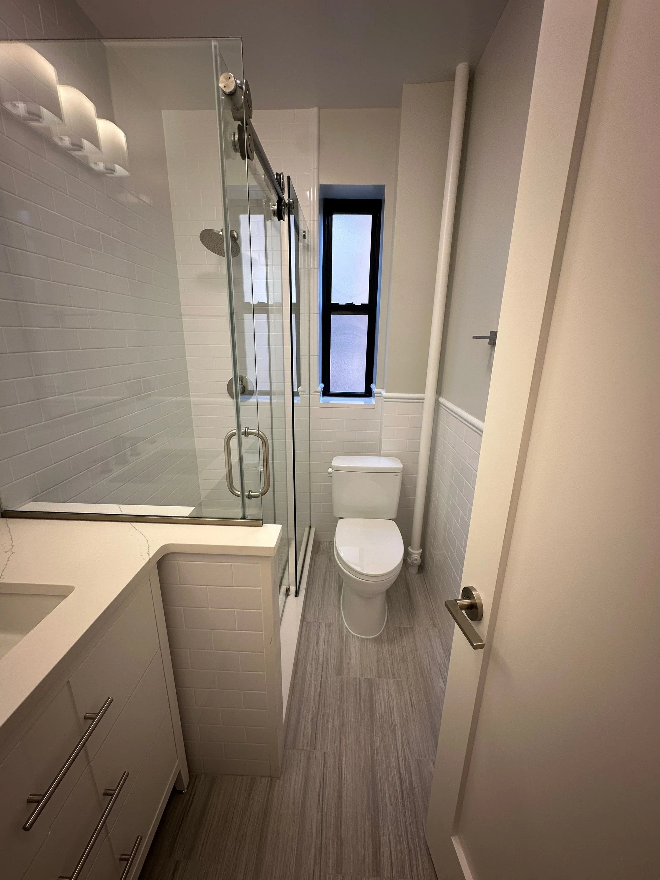 Small modern bathroom with a glass shower enclosure, white tiled walls, a small window, a white toilet, and a beige vanity with a sink.
