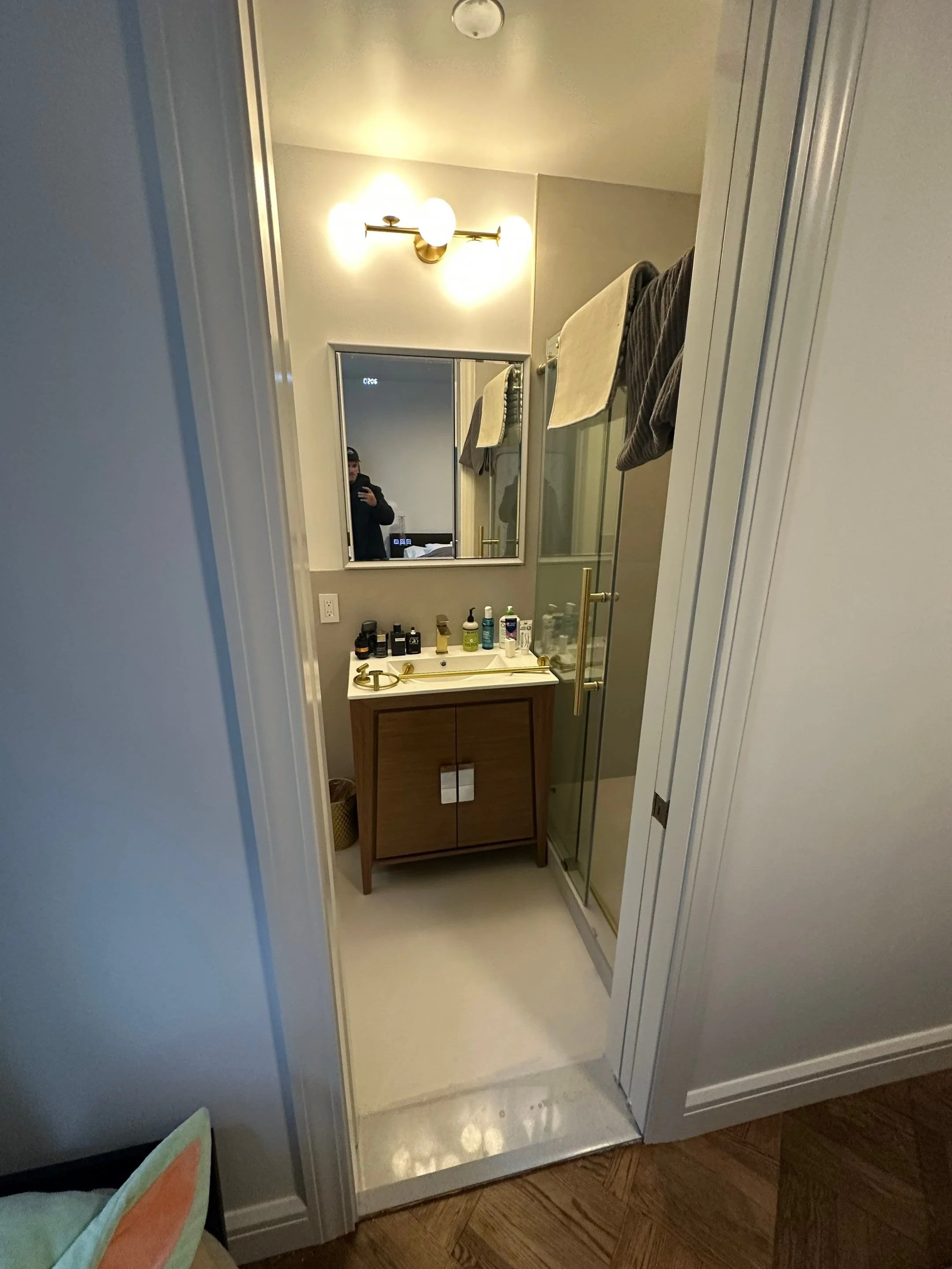 A small bathroom with a wooden vanity, gold fixtures, a mirror, and towels hanging on a rack above the shower, seen through a doorway.