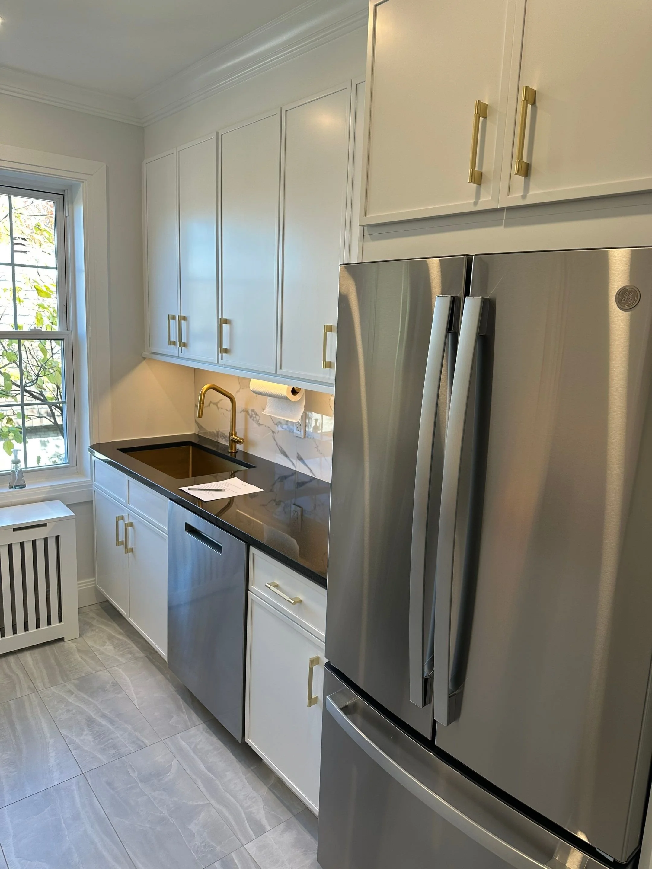Kitchen with white cabinets, black countertop, gold hardware, stainless steel refrigerator, and a black sink with a gold faucet near a window.