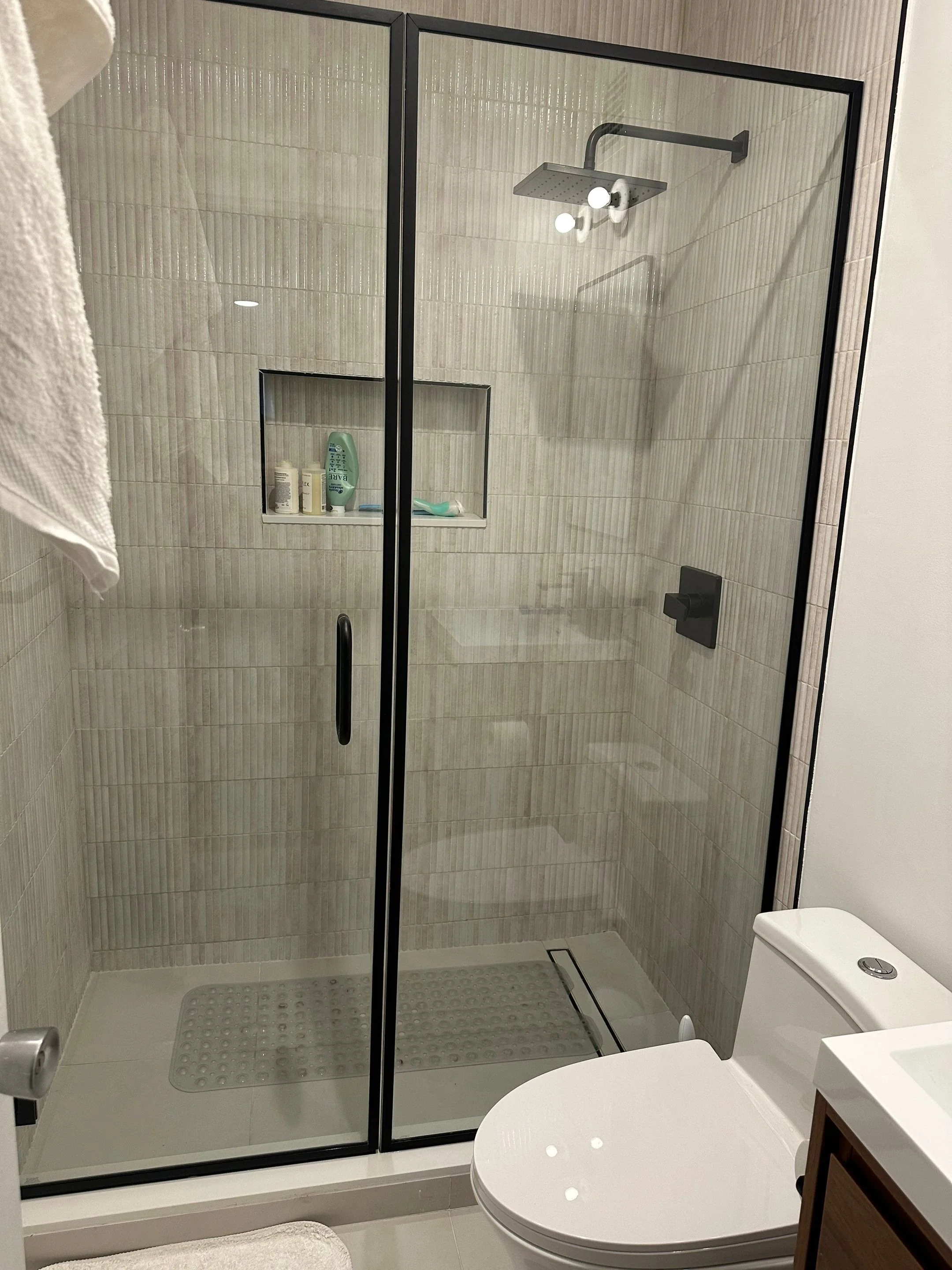 Bathroom with a glass shower enclosure, a built-in shower niche with toiletries, a showerhead, and a toilet next to a small vanity.