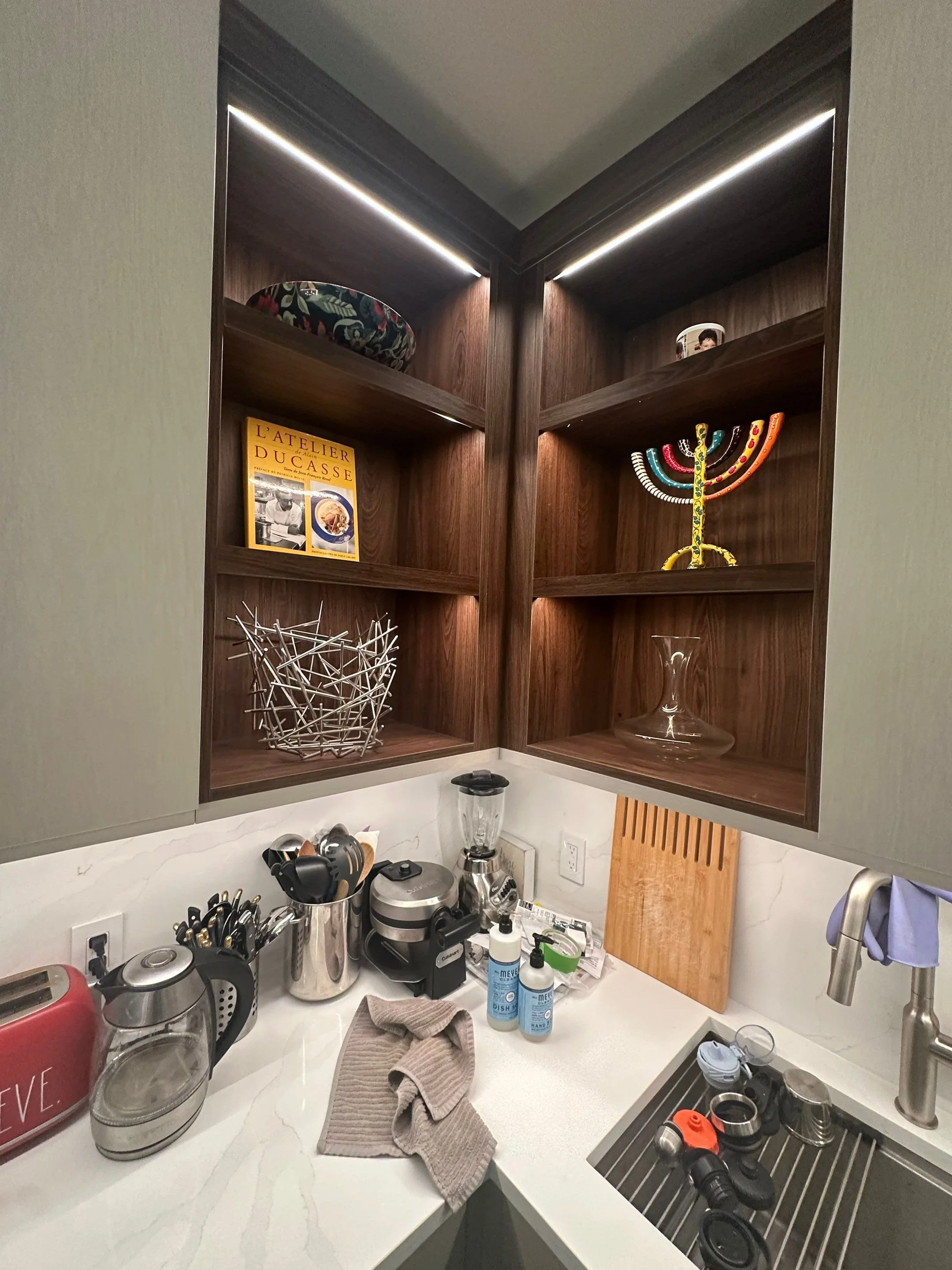 Kitchen corner with upper dark wood cabinets and open shelves, some with decorative items like a menorah, a bowl, and a book, and a kitchen counter below with appliances, utensils, and cleaning supplies.