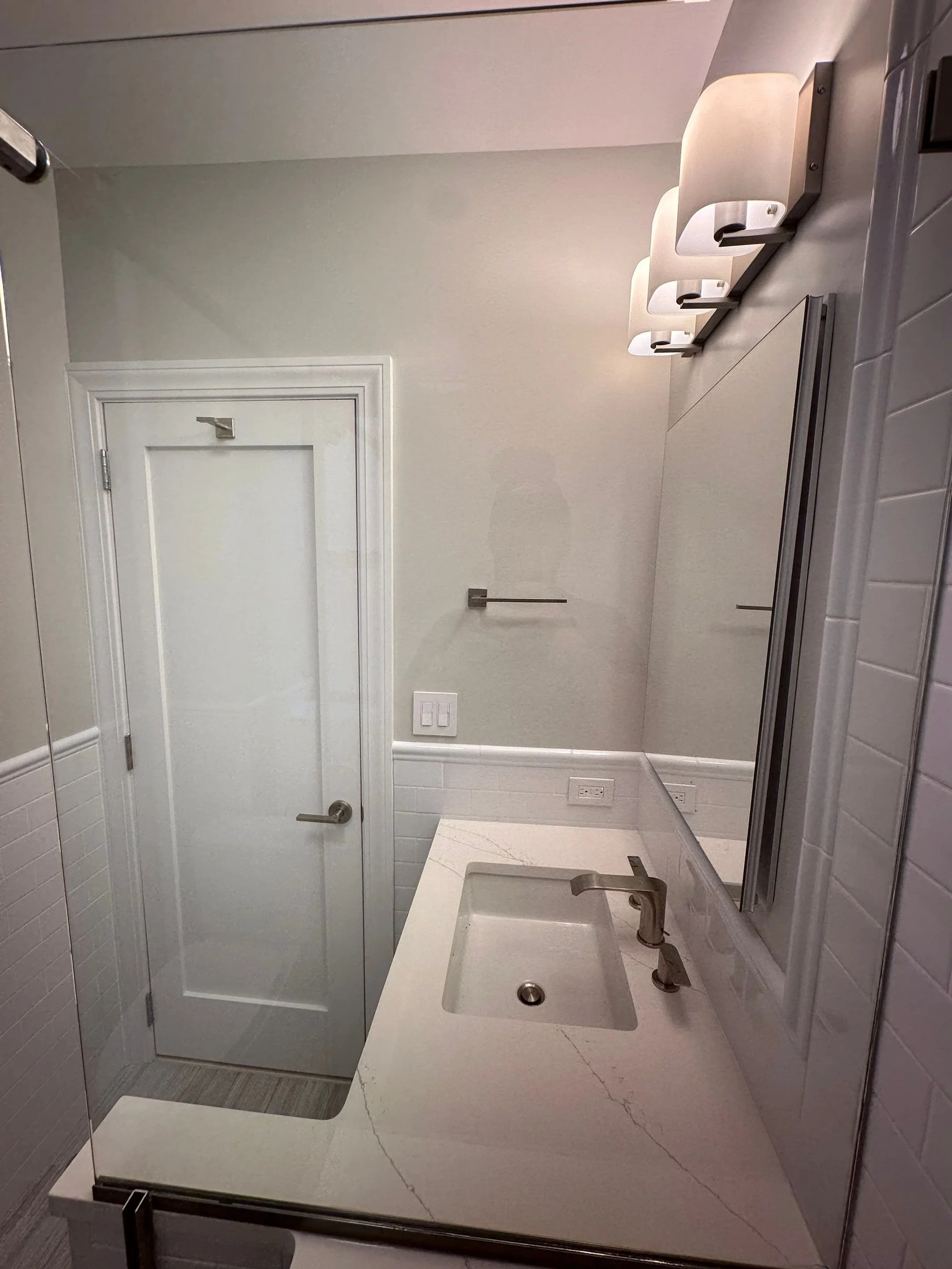 A bathroom with a white door, a marble countertop with an integrated sink, a modern faucet, a large mirror, three white wall-mounted light fixtures, and a towel bar on the wall.