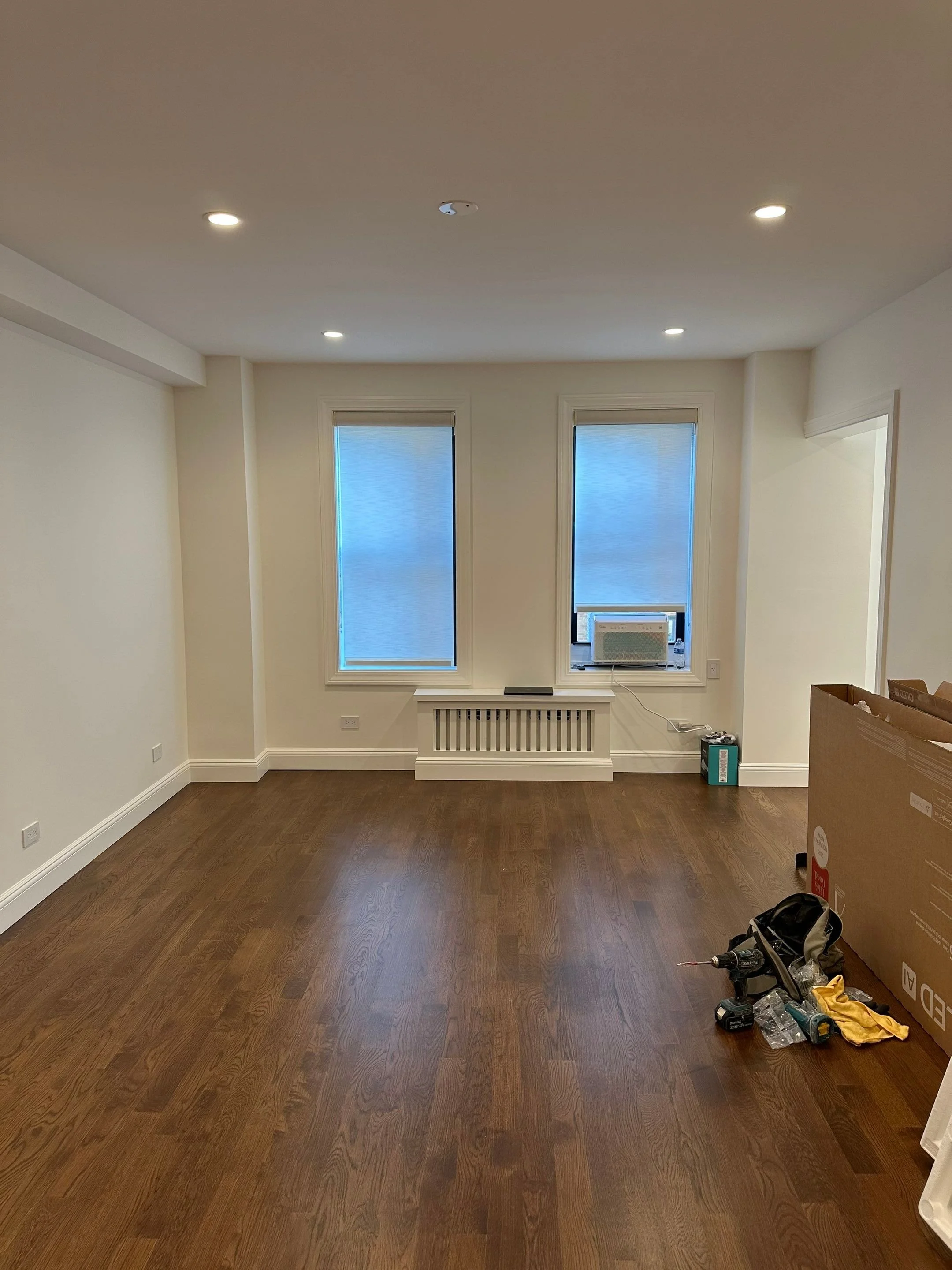 Empty room with hardwood floors, two windows with blinds, a radiator cover beneath the windows, an air conditioner in one window, and some tools and a box in the corner, indicating ongoing interior work.