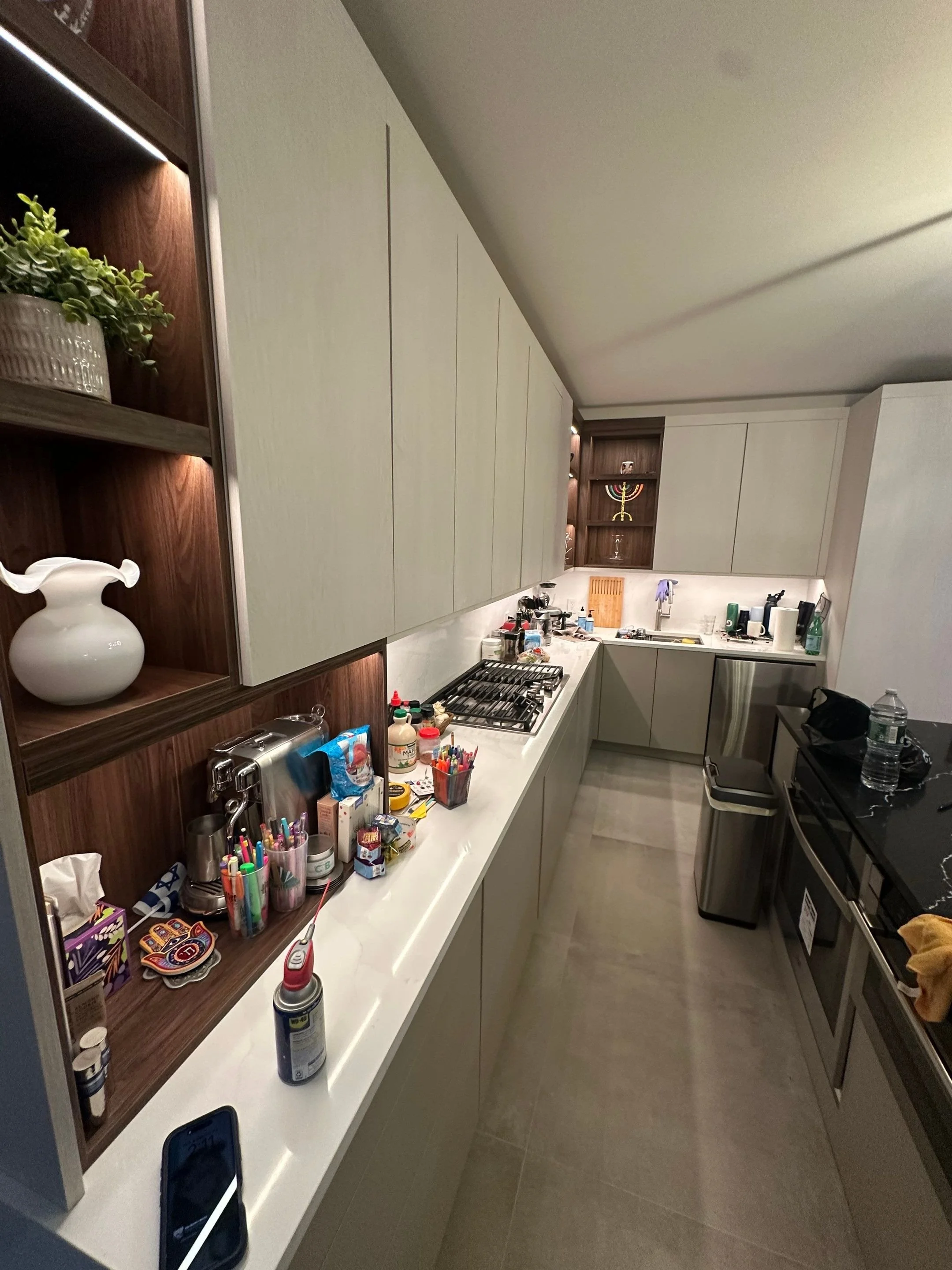 Modern kitchen with white and wood cabinetry, cluttered countertop with various items, gas stove, small sink, and appliances, plants and decorative items on open shelves.