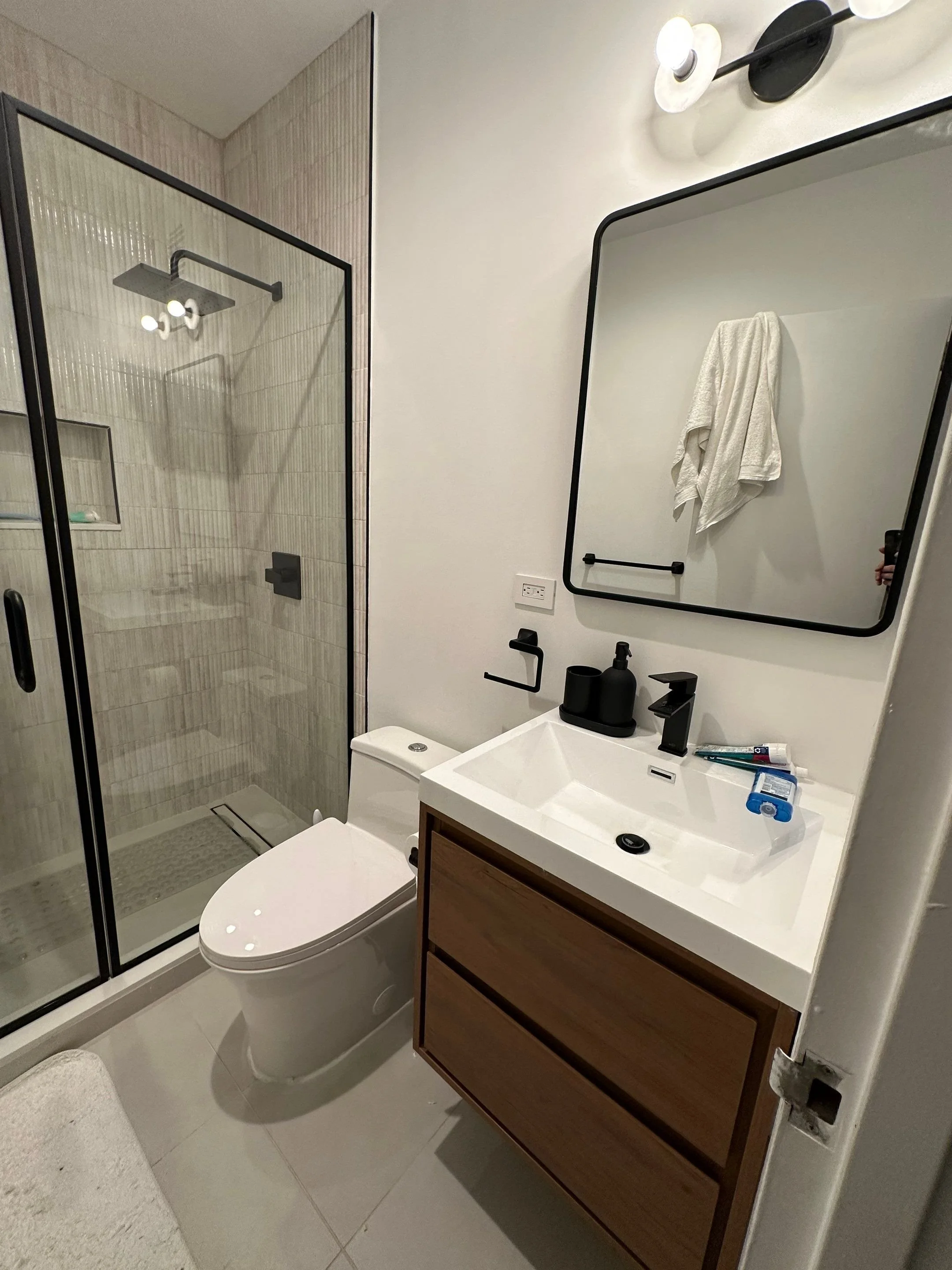 Modern bathroom with walk-in shower, white toilet, and wooden vanity with white sink, black fixtures, and a large mirror. Towel hanging on the wall, toiletries on the sink.