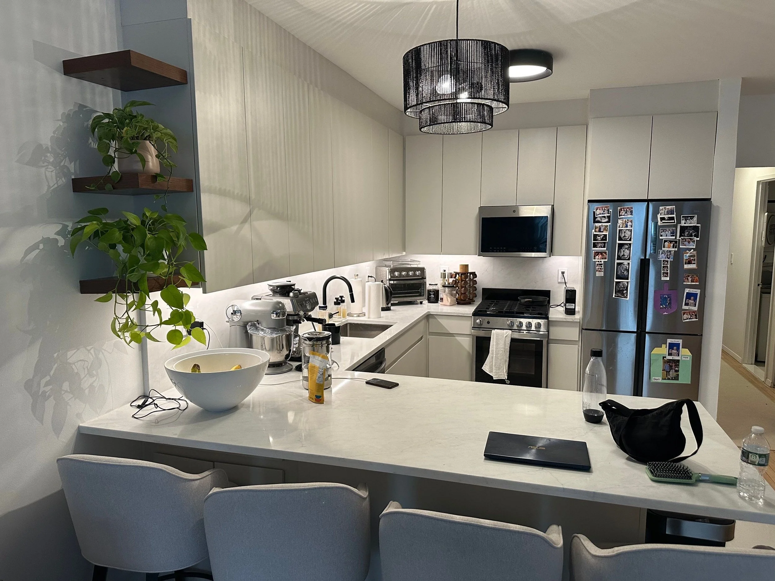 Modern kitchen with white countertops, beige cabinets, and stainless steel appliances. There are potted plants on wall shelves and a black pendant light fixture hanging from the ceiling. The kitchen island has various items including a laptop, water bottles, a hairbrush, and a black bag. The refrigerator door has numerous photos and magnets.