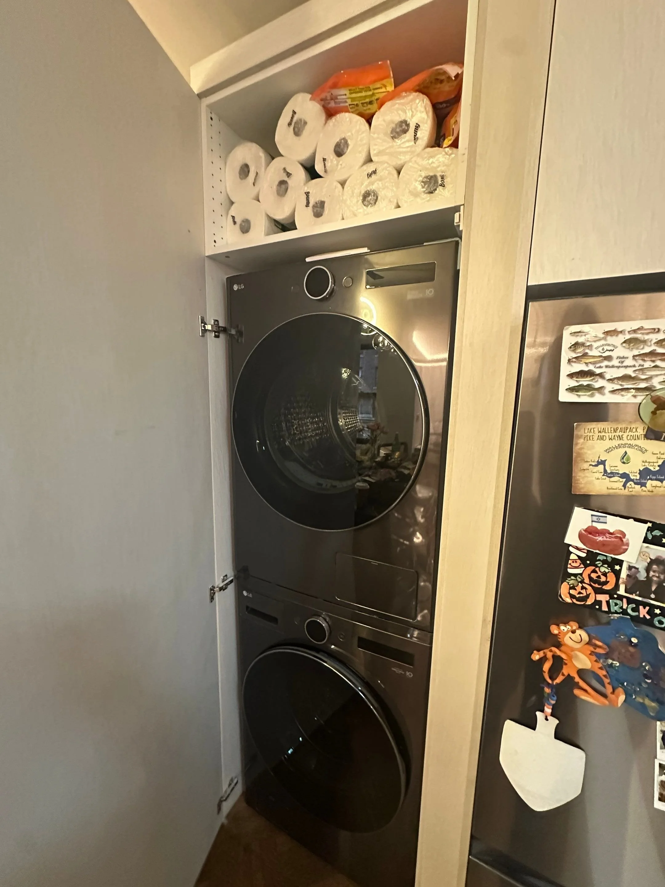 A laundry room with stacked black washer and dryer, a wooden countertop, and a shelf above holding toilet paper rolls and snack items.
