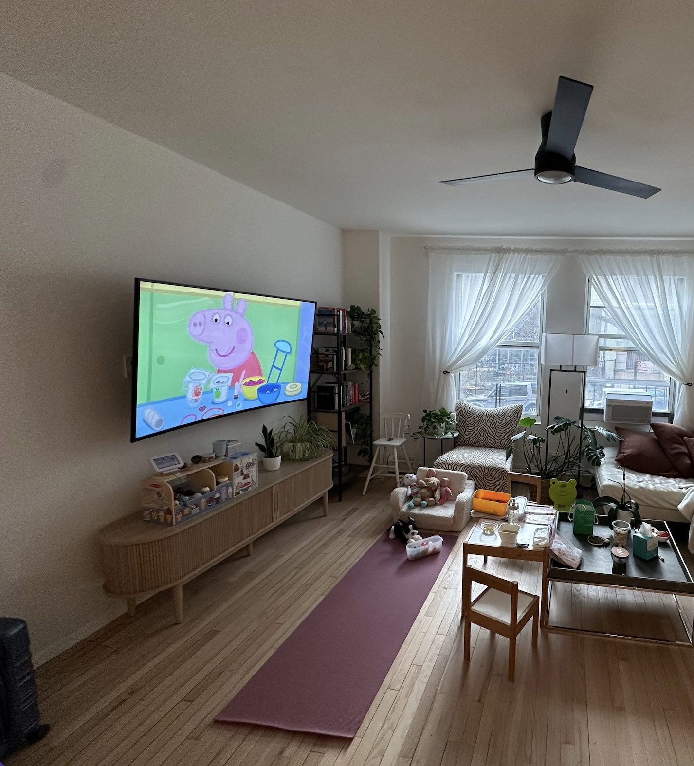 Living room with a wall-mounted TV displaying Peppa Pig, a pink pig. The room has a wooden floor, a pink yoga mat, a small sofa with stuffed animals, a wooden coffee table with toys and cups, and large windows with white curtains.