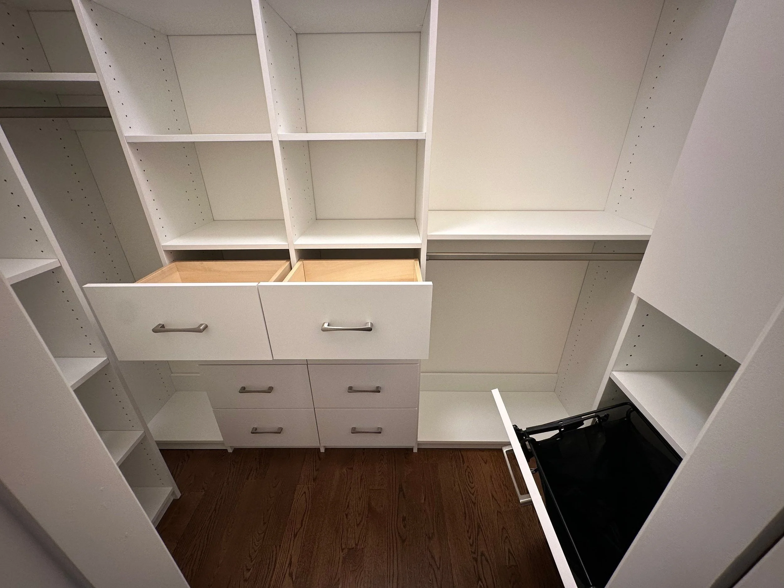 Empty white kitchen cabinets with open drawers, shelves, and a black stove in a small kitchen with wooden flooring.