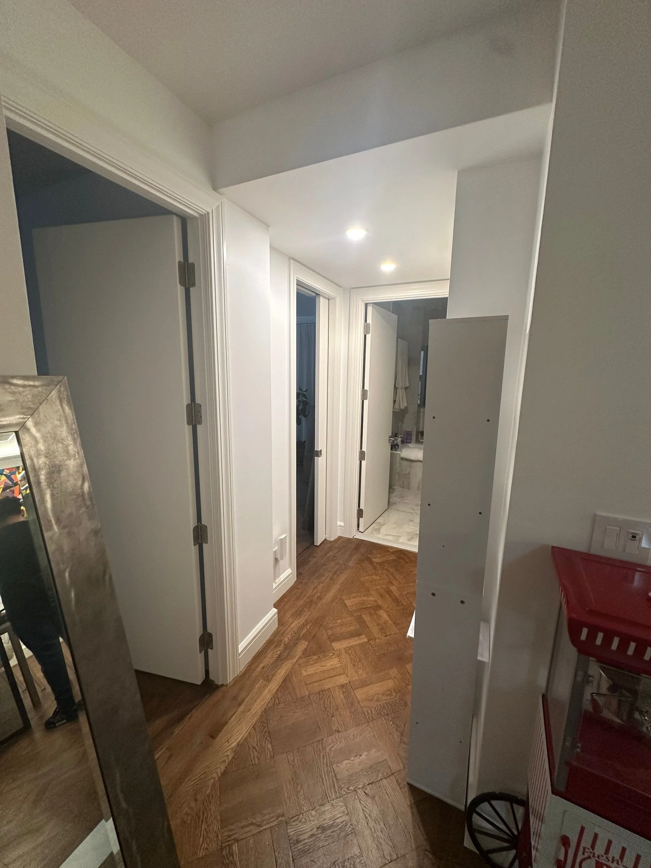 A hallway with wooden parquet flooring, white walls, and multiple doors leading to rooms; there is a mirror reflecting part of a person and furniture.