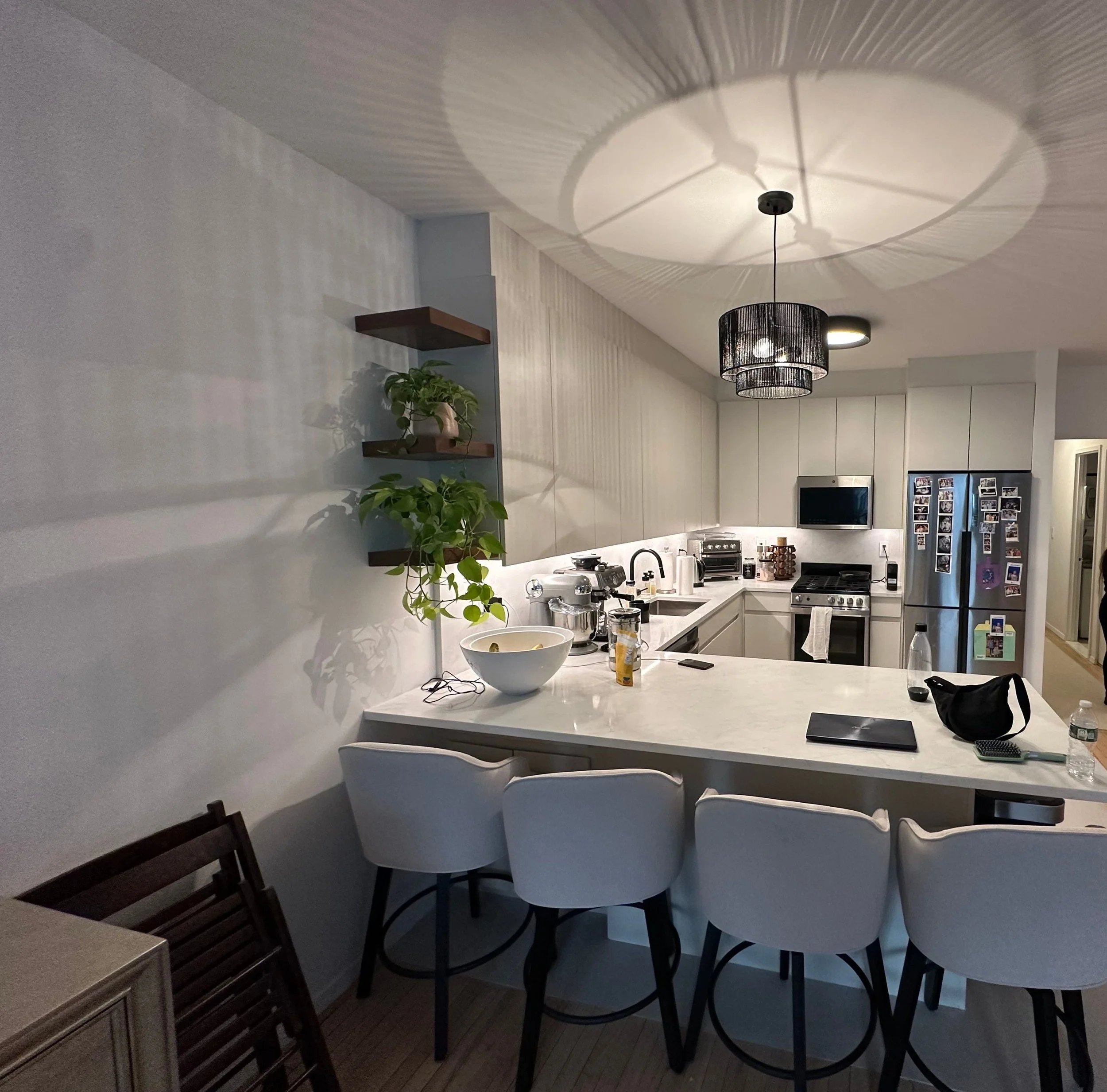 Modern kitchen with white cabinets, stainless steel appliances, and a marble island with bar stools. Potted plants are on floating shelves, and a black ceiling light casts shadows on the ceiling.