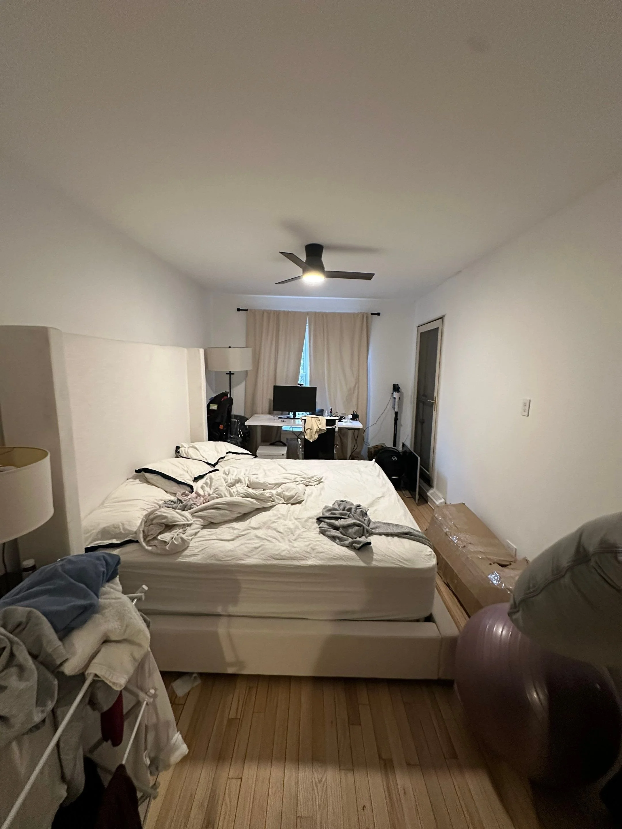Unfinished bedroom with unmade bed, laundry basket, exercise ball, cluttered desk, and various items scattered around.