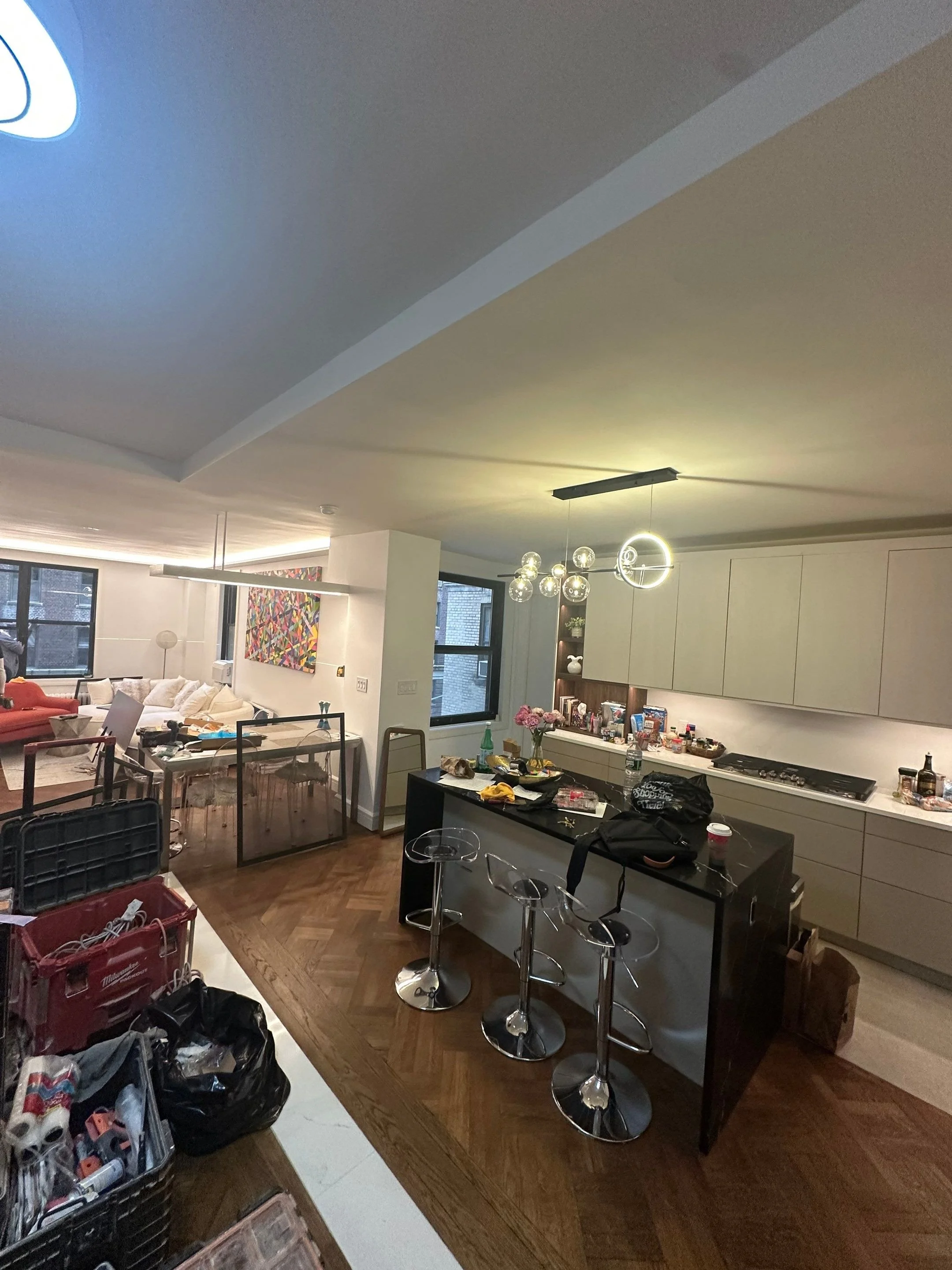 Messy apartment kitchen with clutter on black island counter, stools, and dirty dishes; living area with sofa and artwork; boxes and supplies on the floor.