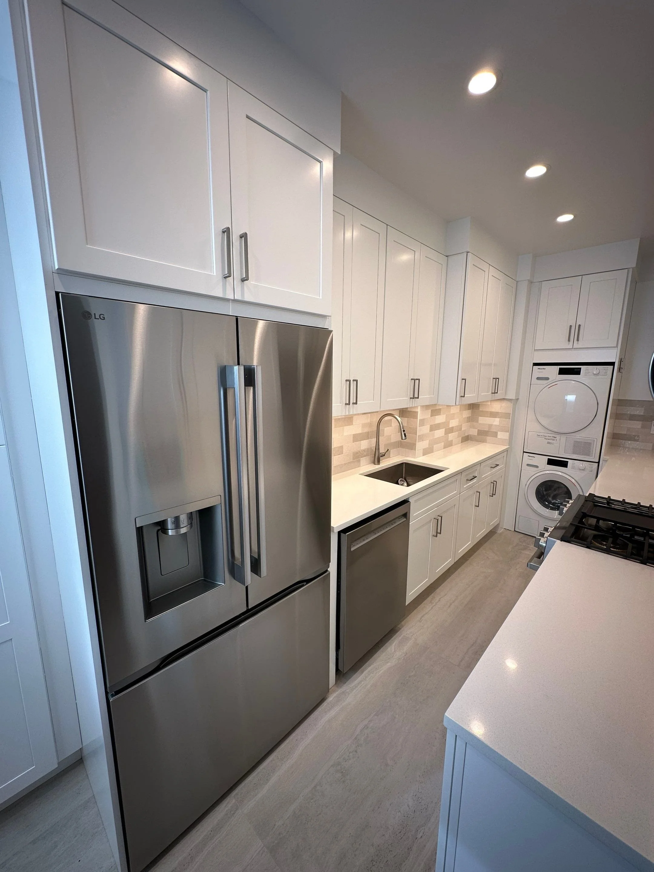 Modern kitchen with white cabinets, stainless steel LG refrigerator, small built-in dishwasher, and stacked washer and dryer in the background.