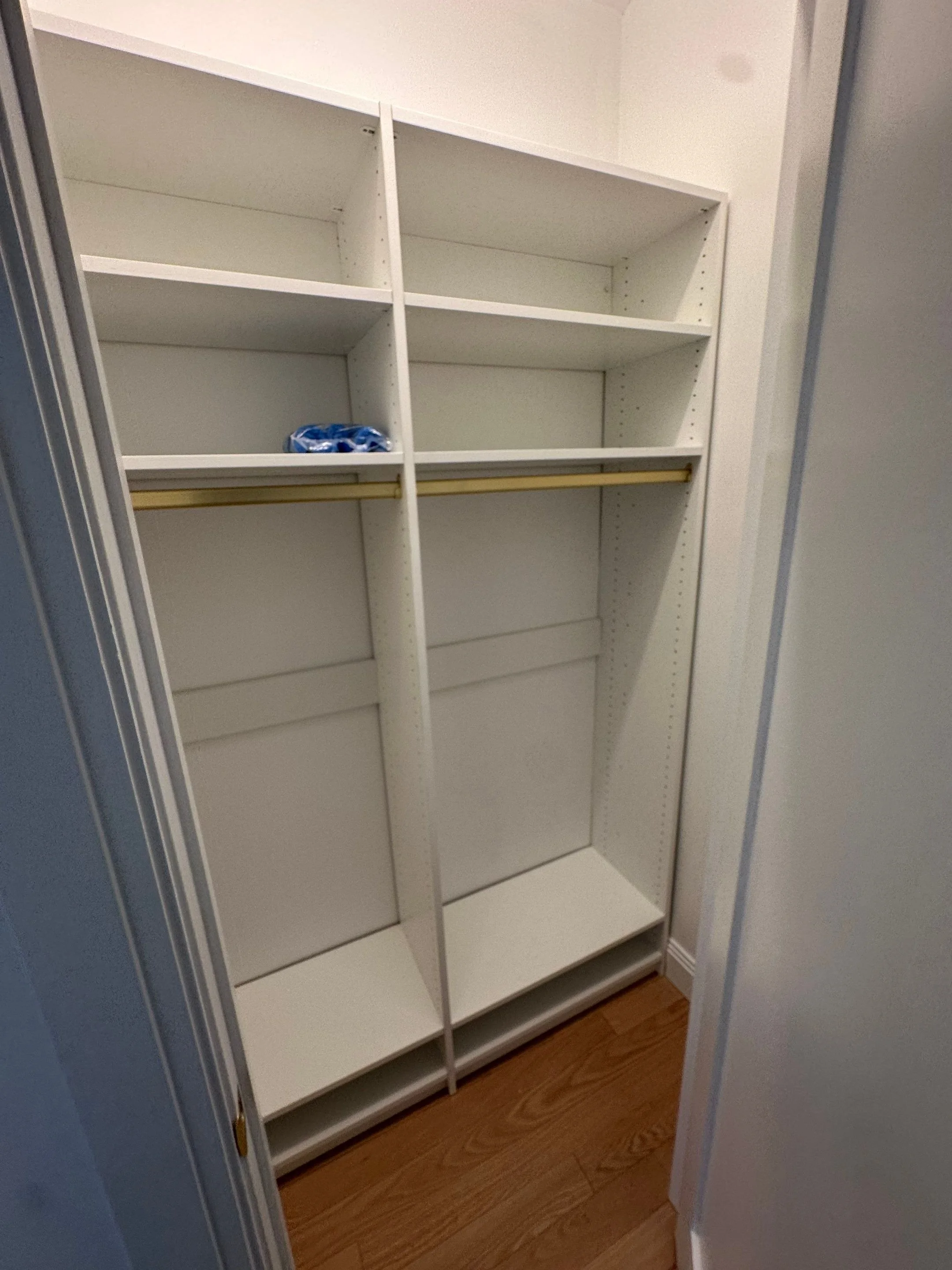 Empty white closet with shelves and a golden clothing rod, located next to a hardwood floor.