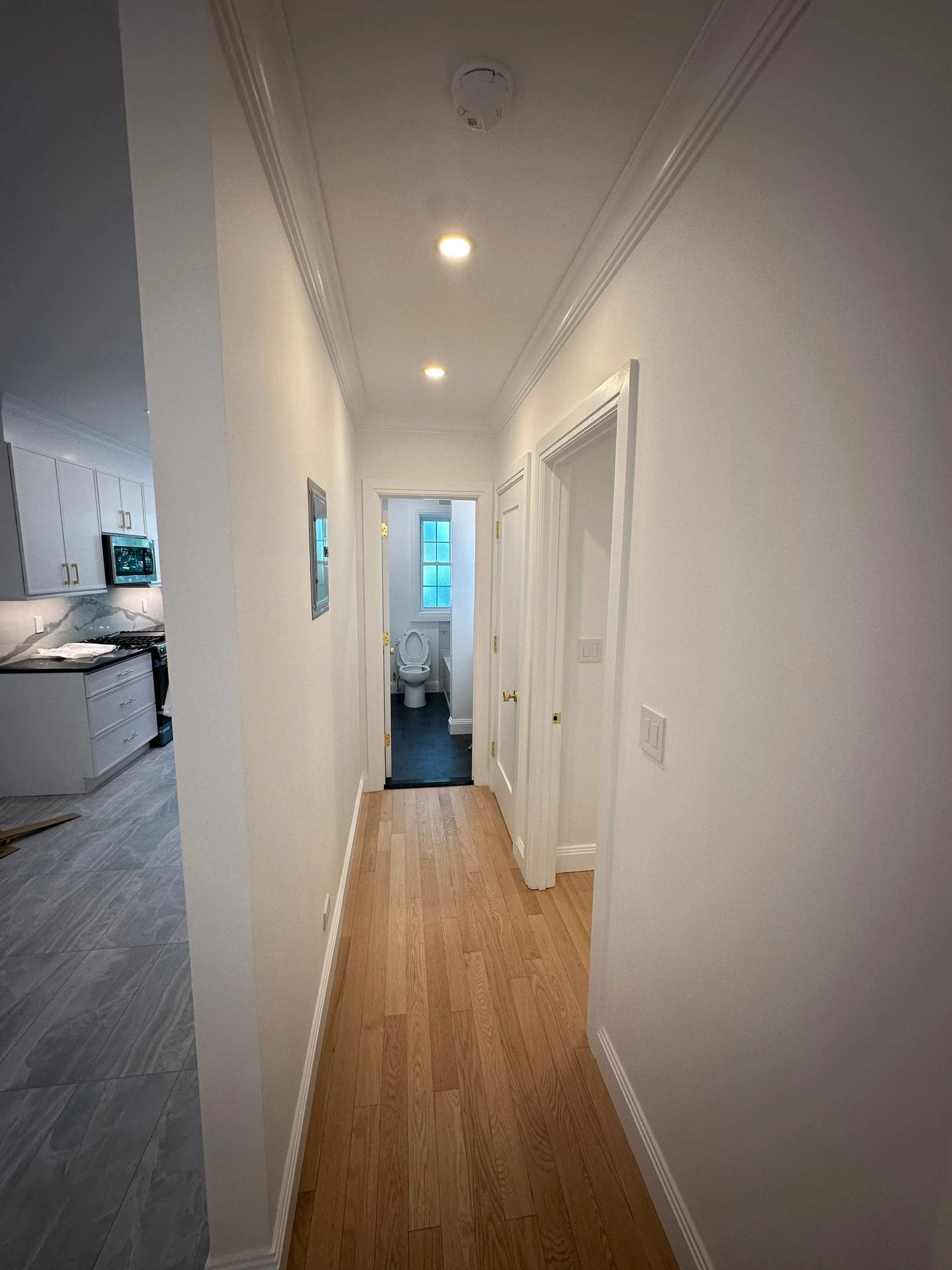 Interior view of a narrow hallway with wooden flooring leading to a bathroom at the end. The hallway has white walls, crown molding, and ceiling lights. Part of a kitchen with gray cabinets and marble countertops is visible on the left.
