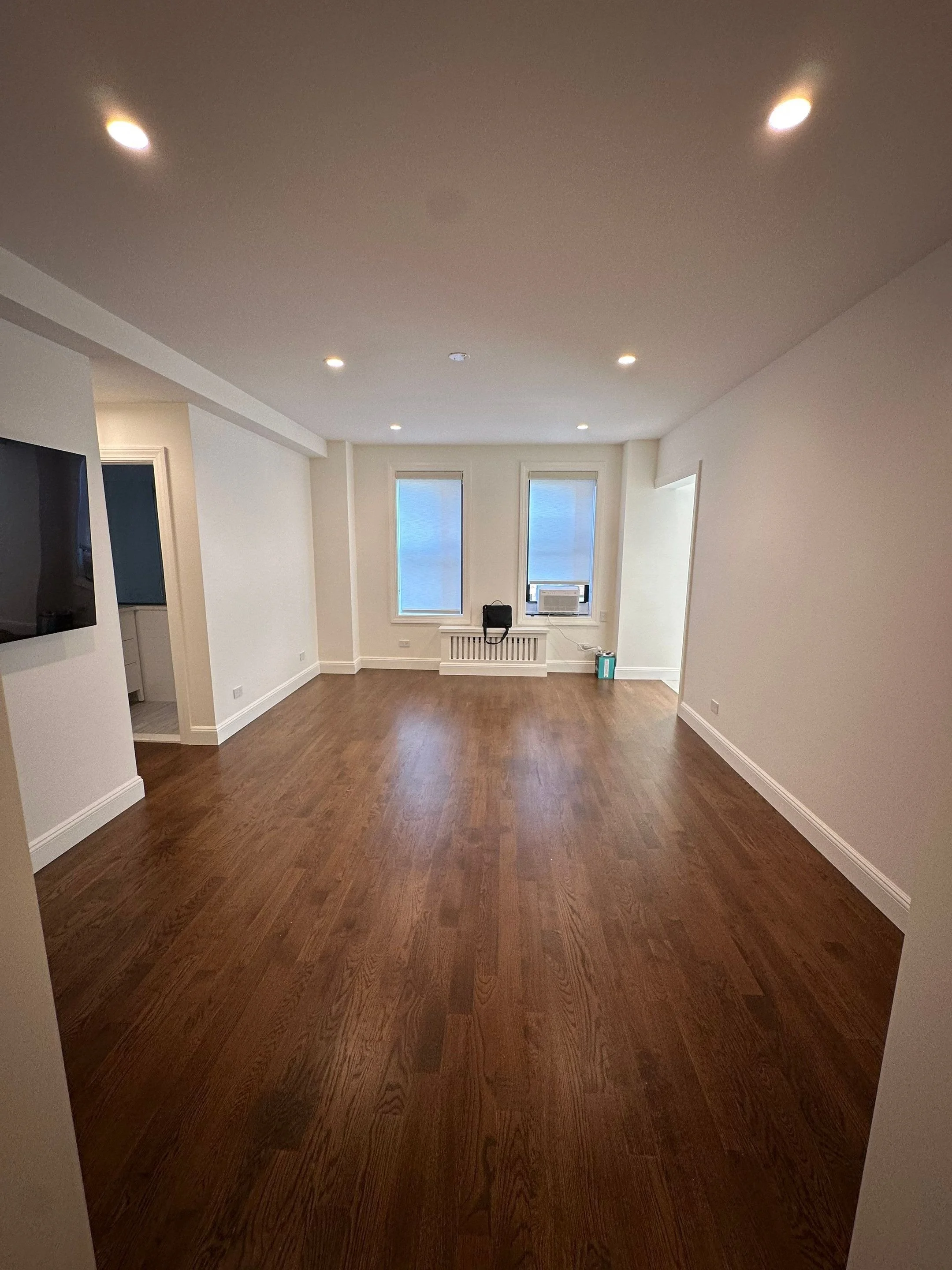 Empty living room with hardwood floors, white walls, recessed ceiling lights, and two windows with blinds.