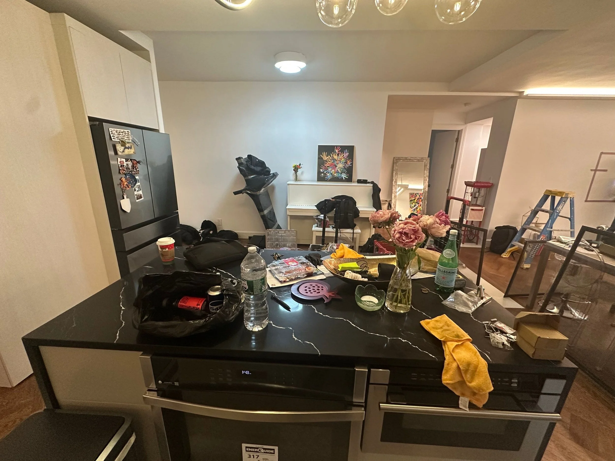 A cluttered kitchen counter with various items including pink flowers in a glass vase, water bottles, a black bag, a yellow cloth, and snack packs, with a background of a white piano, a mirror, and a ladder in a brightly lit room.
