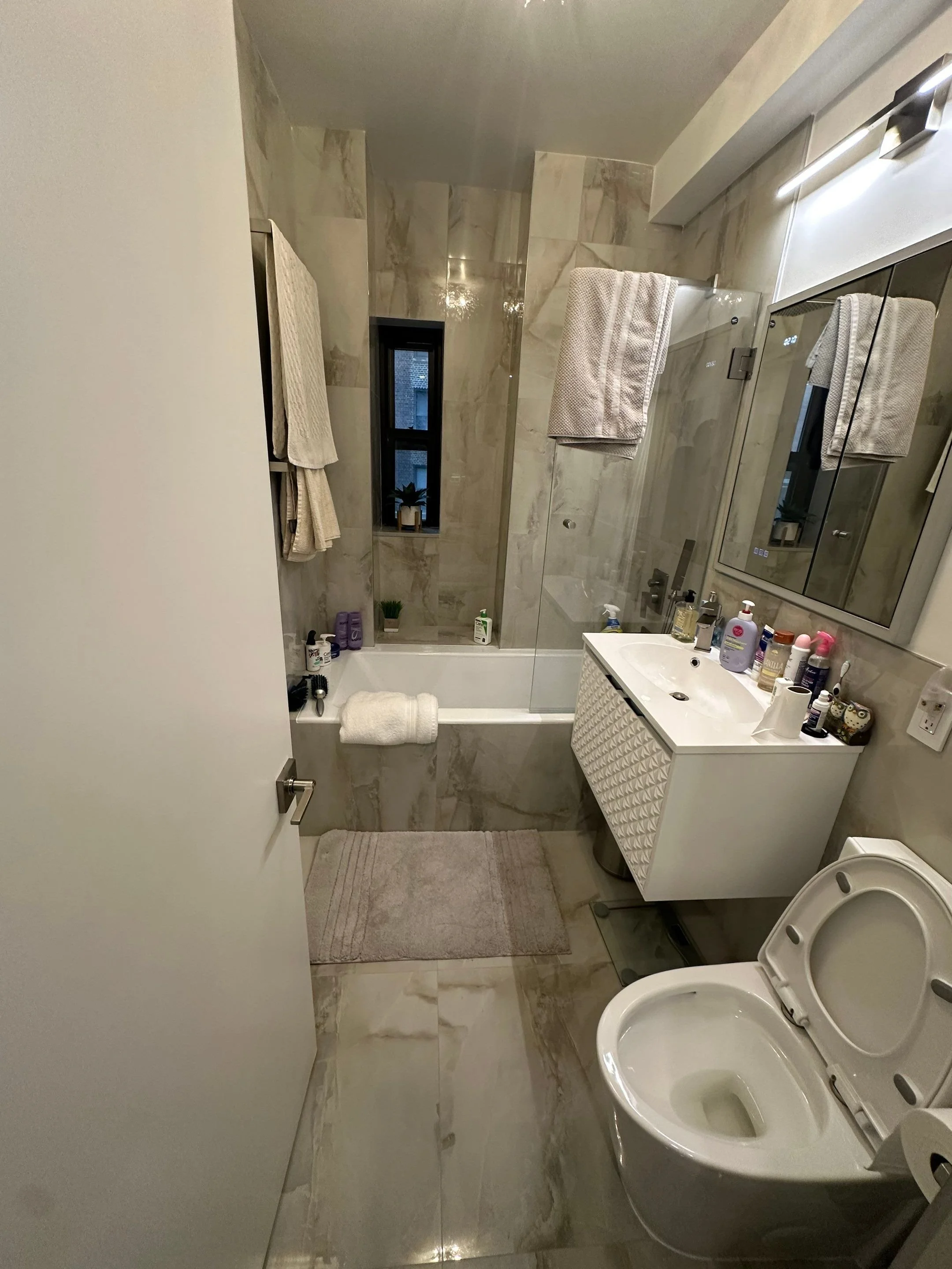 Small modern bathroom with beige marble walls and floor, a bathtub with a shower, a white vanity with a mirror, and a toilet.