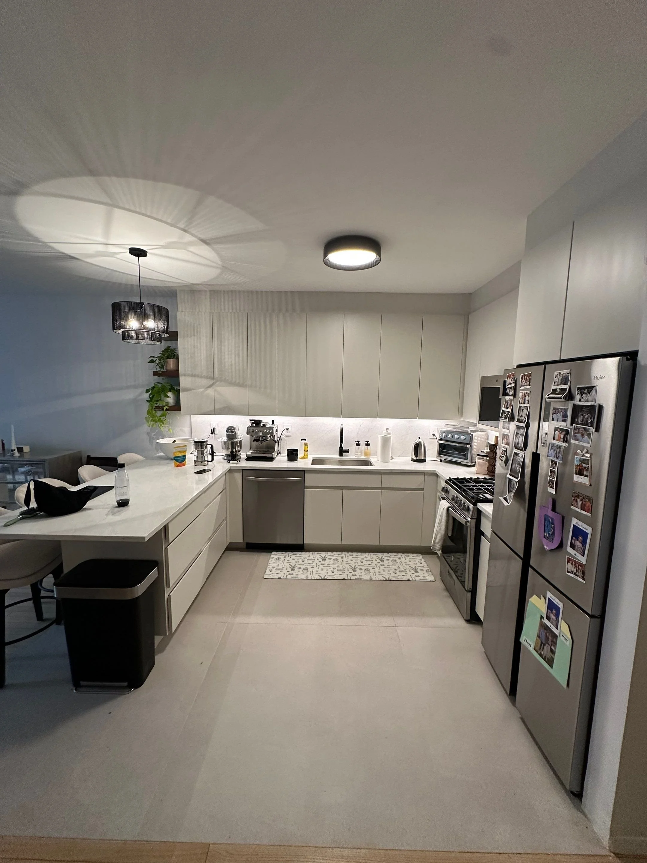 Modern kitchen with white cabinetry, stainless steel appliances, a fridge covered with photos, and a marble white countertop with various kitchen items. There is a hanging light fixture, and a small rug on the floor in front of the sink.