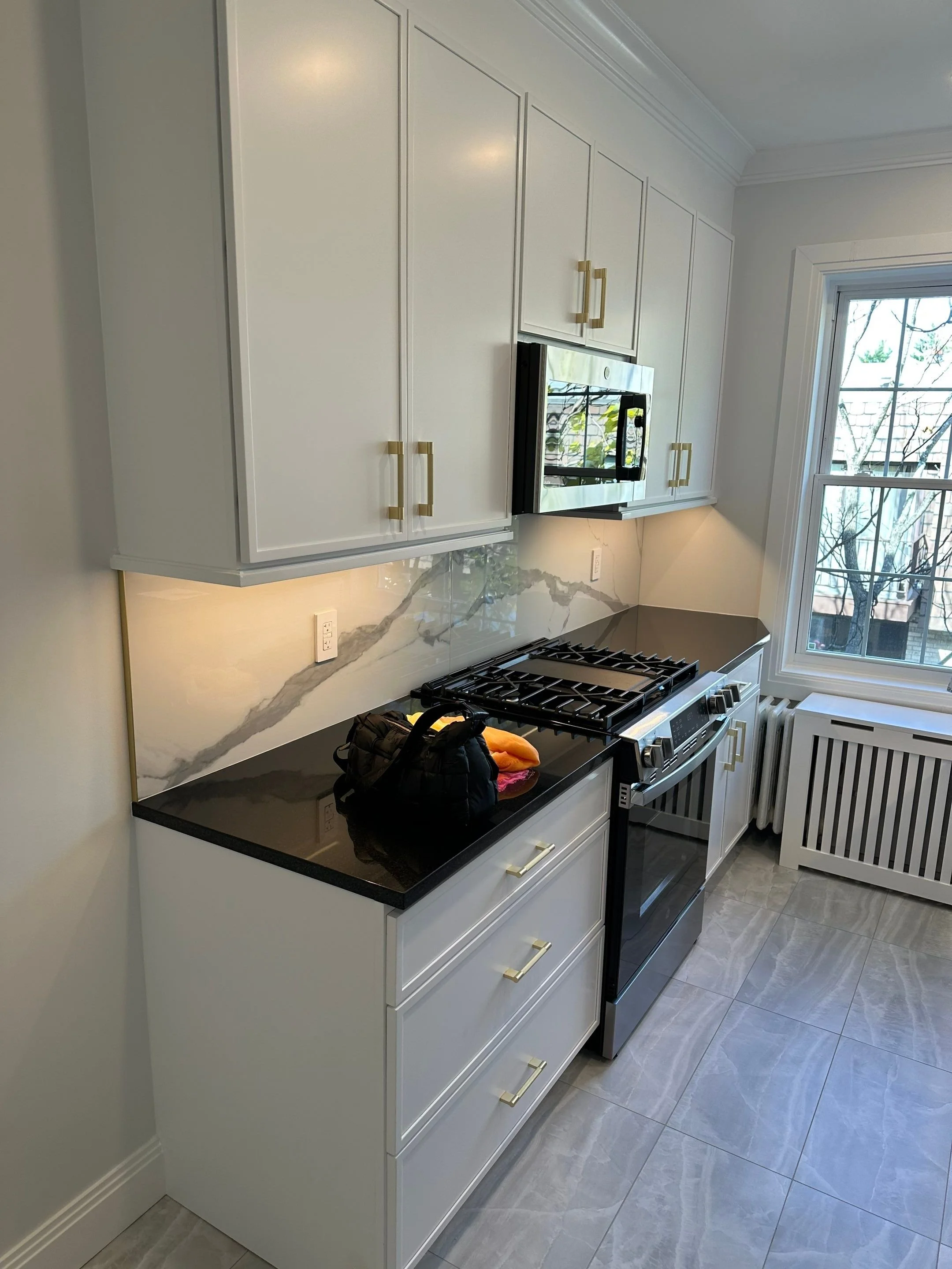 Modern kitchen with white cabinetry, black countertop, stainless steel microwave oven above stove, and a window showing trees outside.