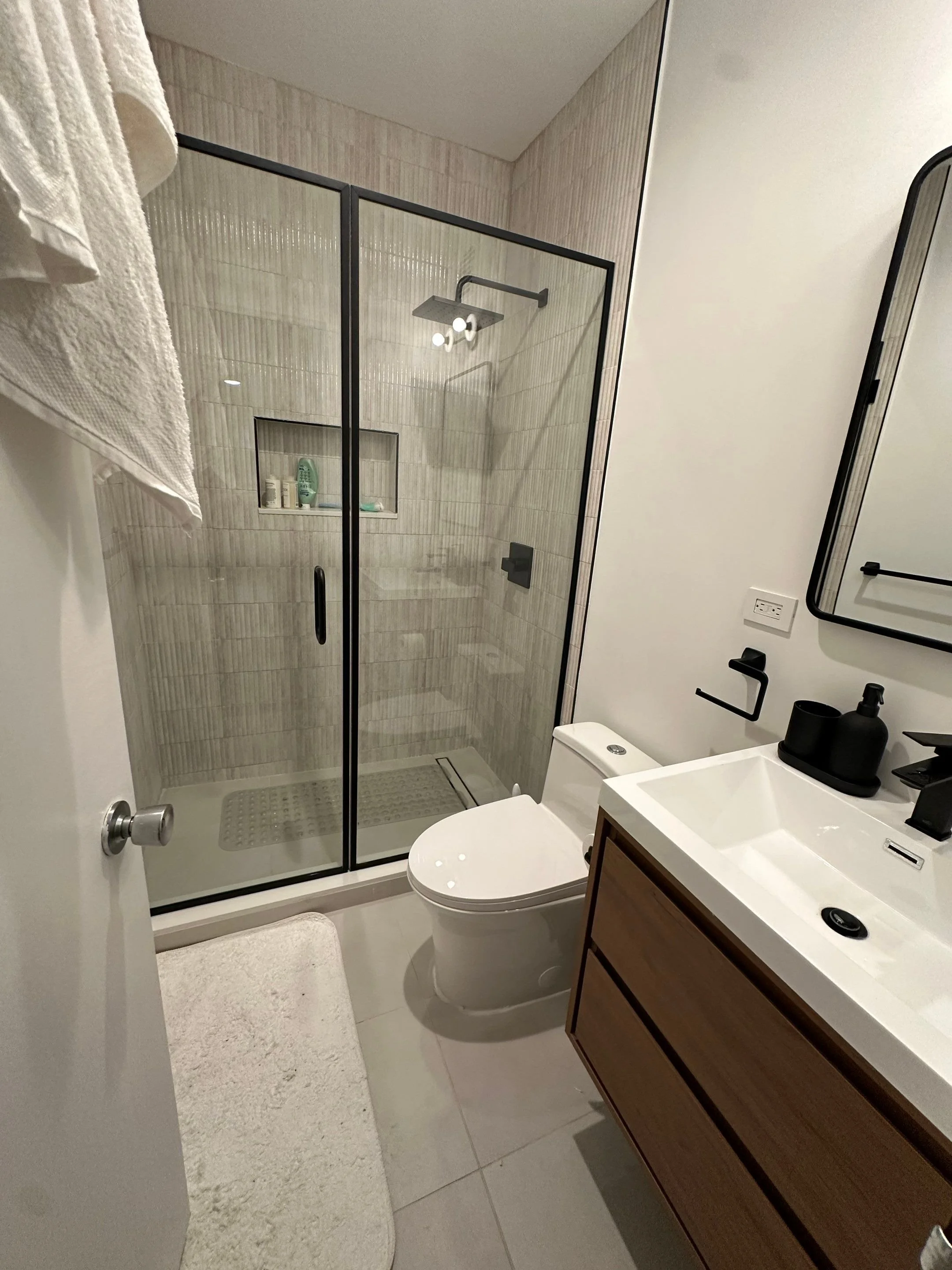 A modern bathroom with a glass shower enclosure, wooden vanity with white sink, and a white toilet. There are towels hanging on the wall and toiletries inside the shower. A mirror and black fixtures are also visible.