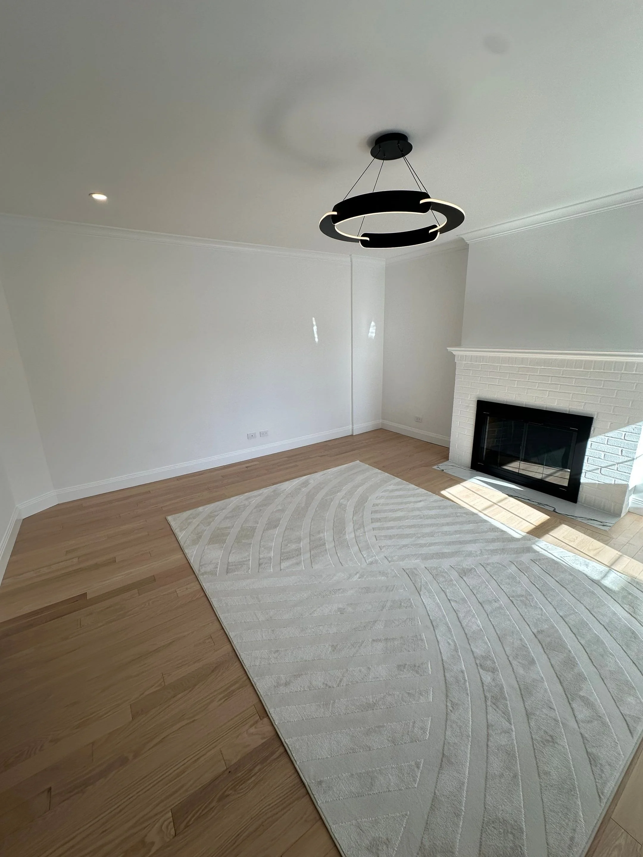Empty living room with white walls, a modern black chandelier, a fireplace, hardwood floors, and a light-colored area rug.