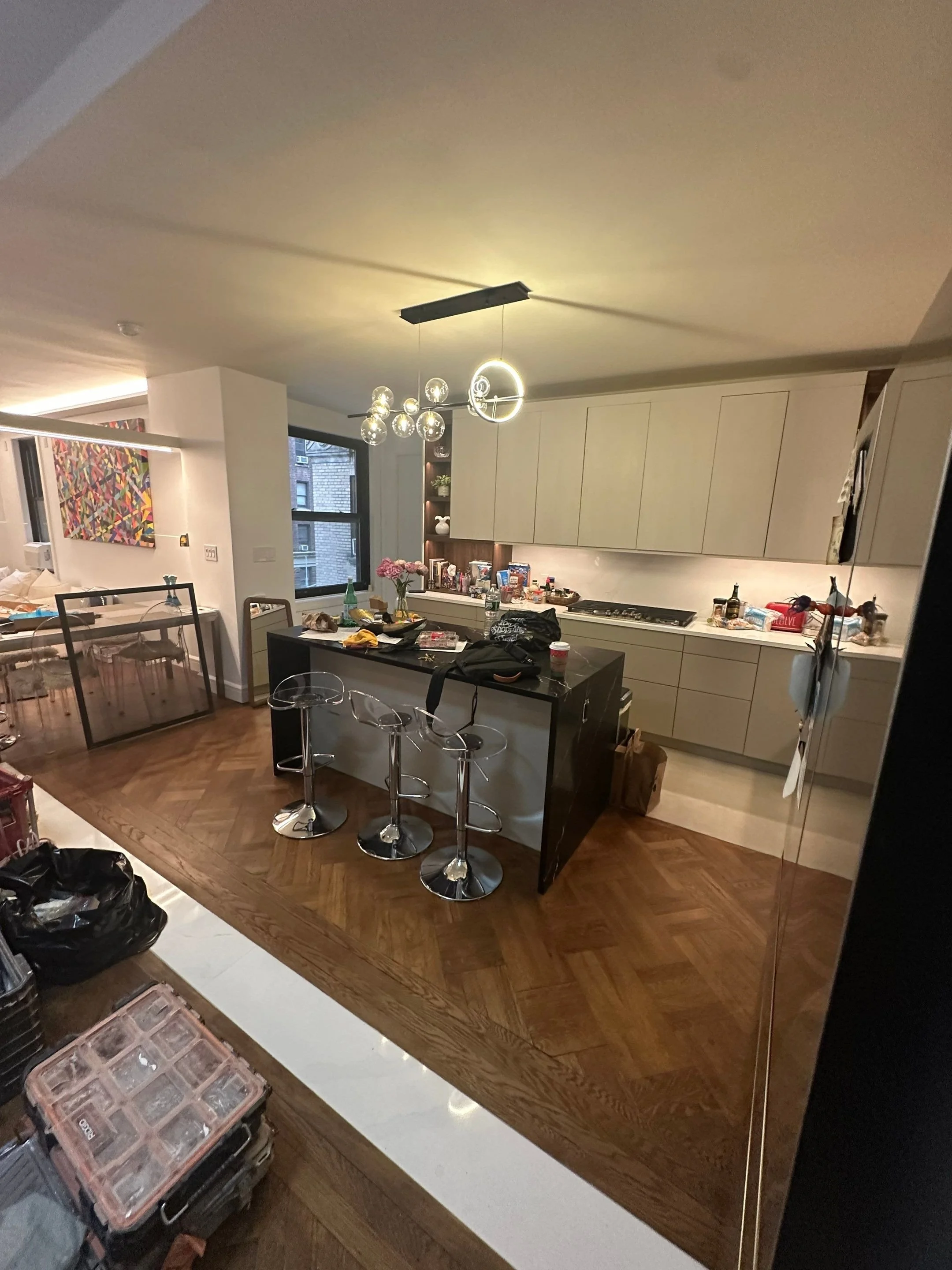 Modern kitchen with a black island, three clear bar stools, beige cabinetry, and a wooden herringbone floor. The countertop has various items including flowers, bottles, and food.