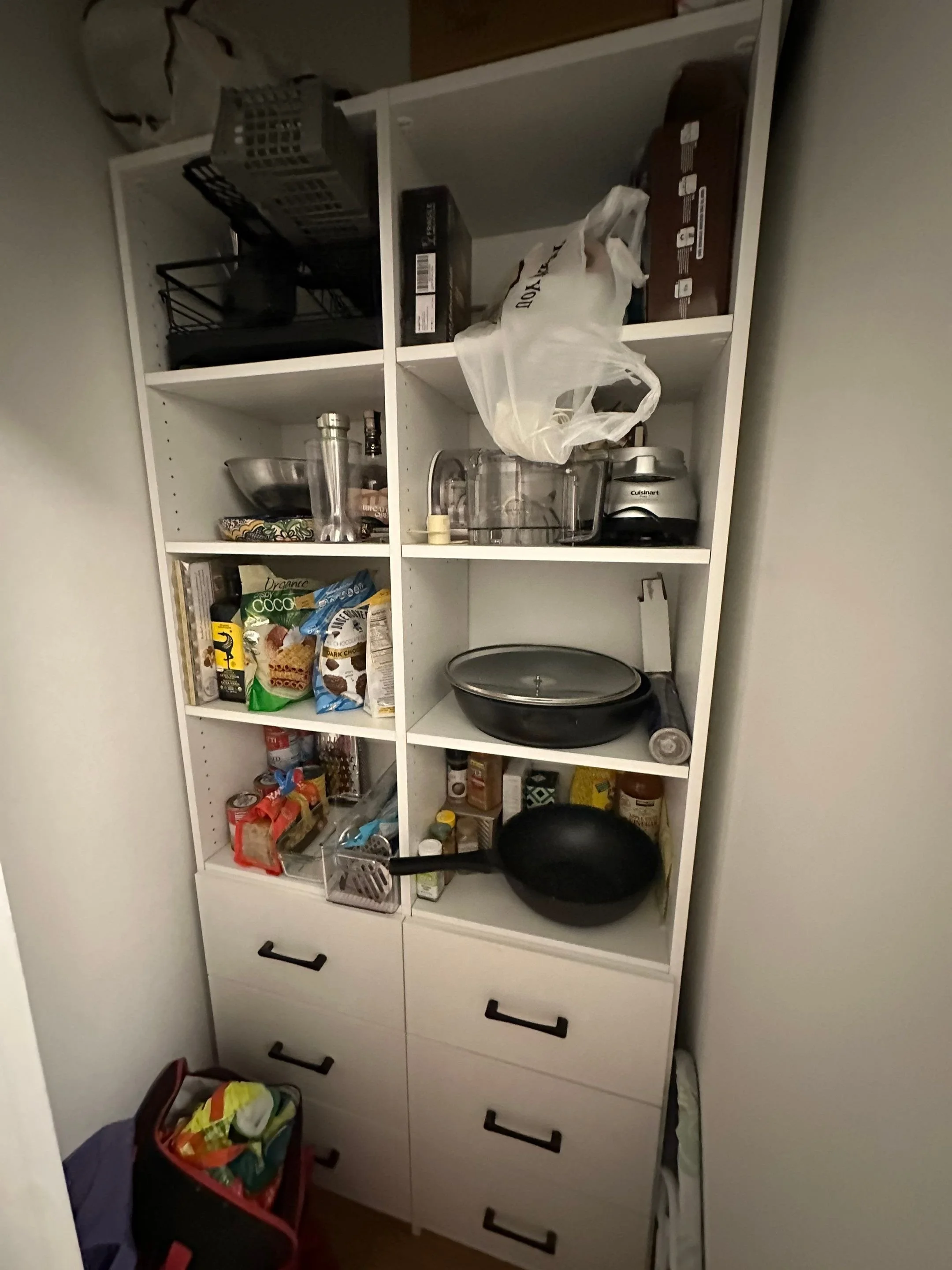 A white kitchen cabinet filled with various kitchen items, including pots, pans, jars, boxes, and plastic bags, with a small trash bin in front.