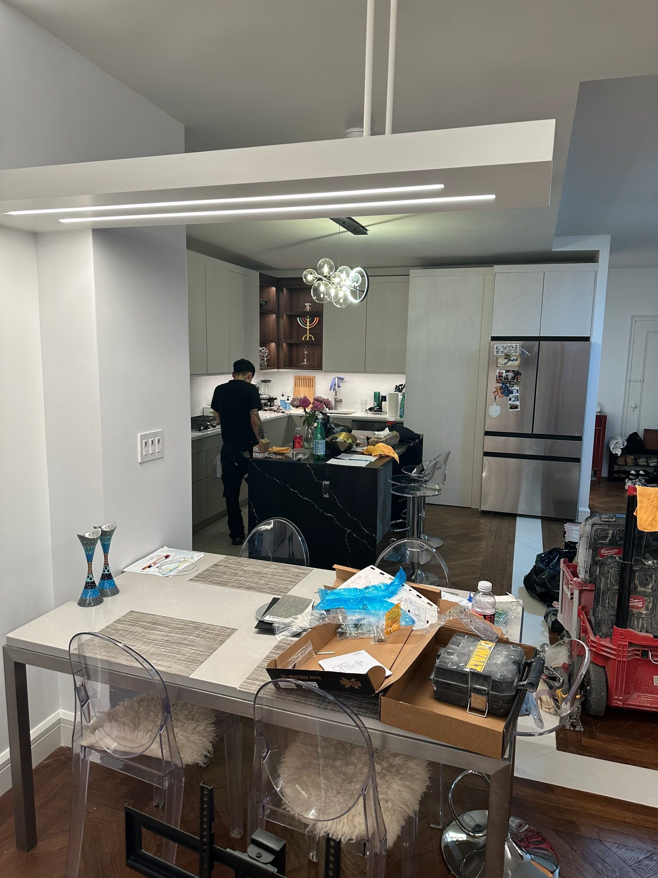 A modern open kitchen with a man working at the counter, a dining table with various items and tools, and construction or renovation supplies scattered around.