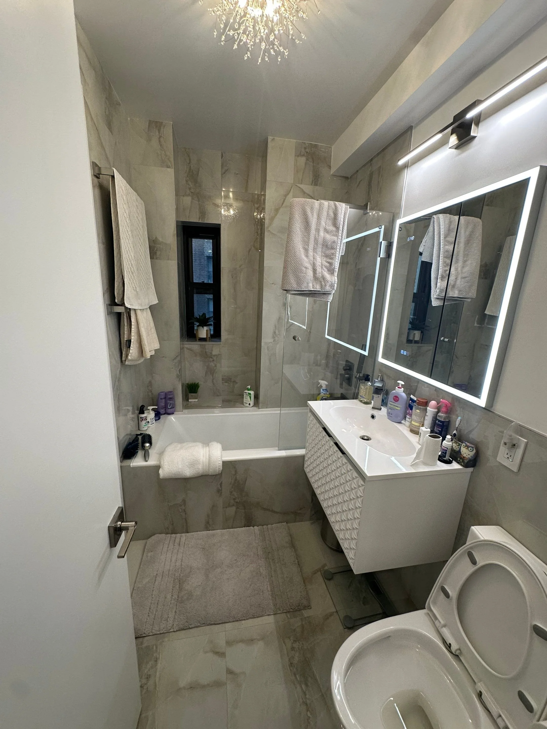 Modern bathroom with marble tiles, a bathtub with a glass shower screen, a small window, a vanity with a mirror and various toiletries, towel racks with towels, a toilet with the lid open, and a gray bath mat.