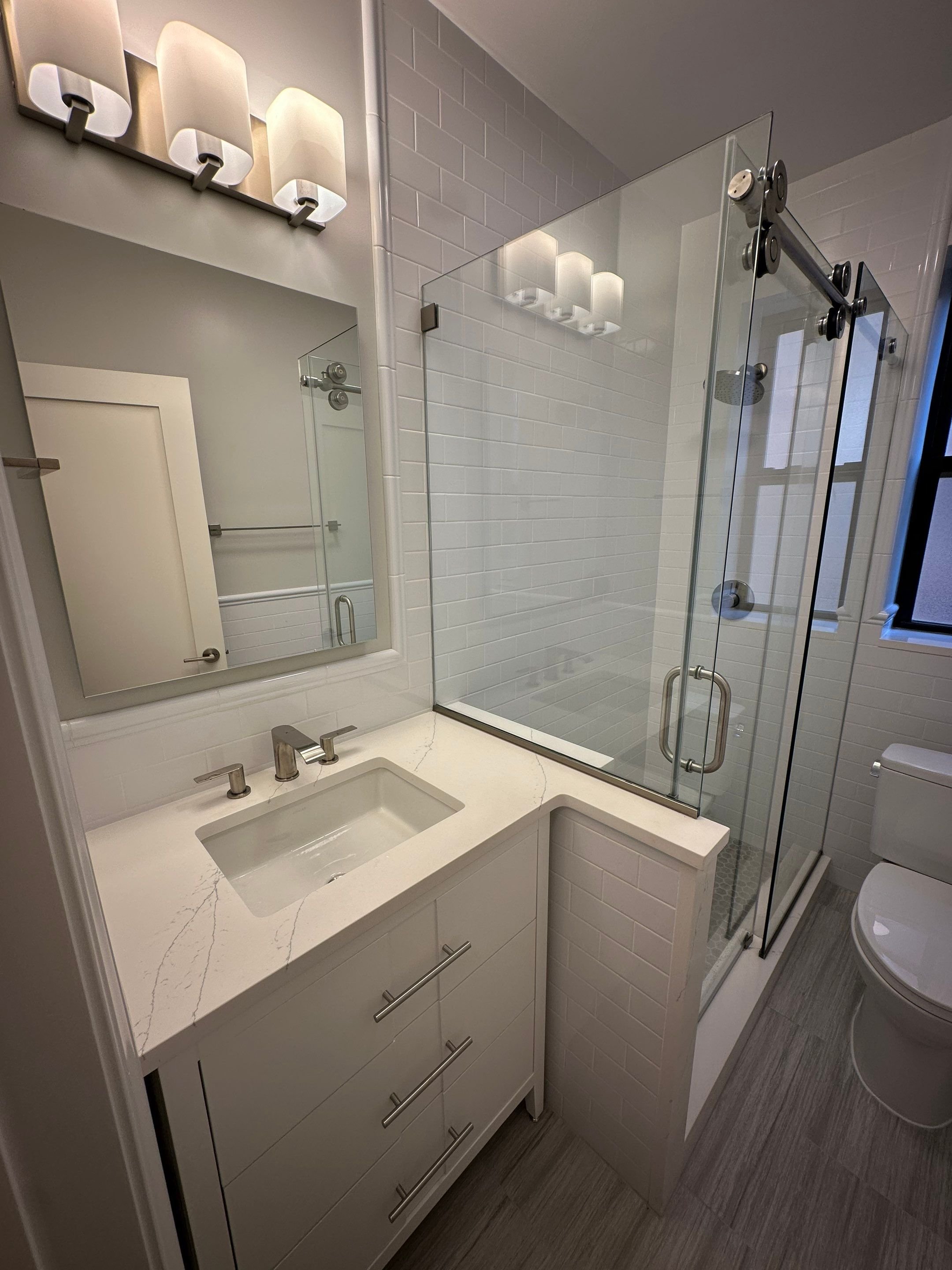 Modern bathroom with white vanity and marble countertop, rectangular sink, brushed nickel fixtures, large mirror, white subway tile walls, glass shower enclosure with sliding door, and toilet near window.