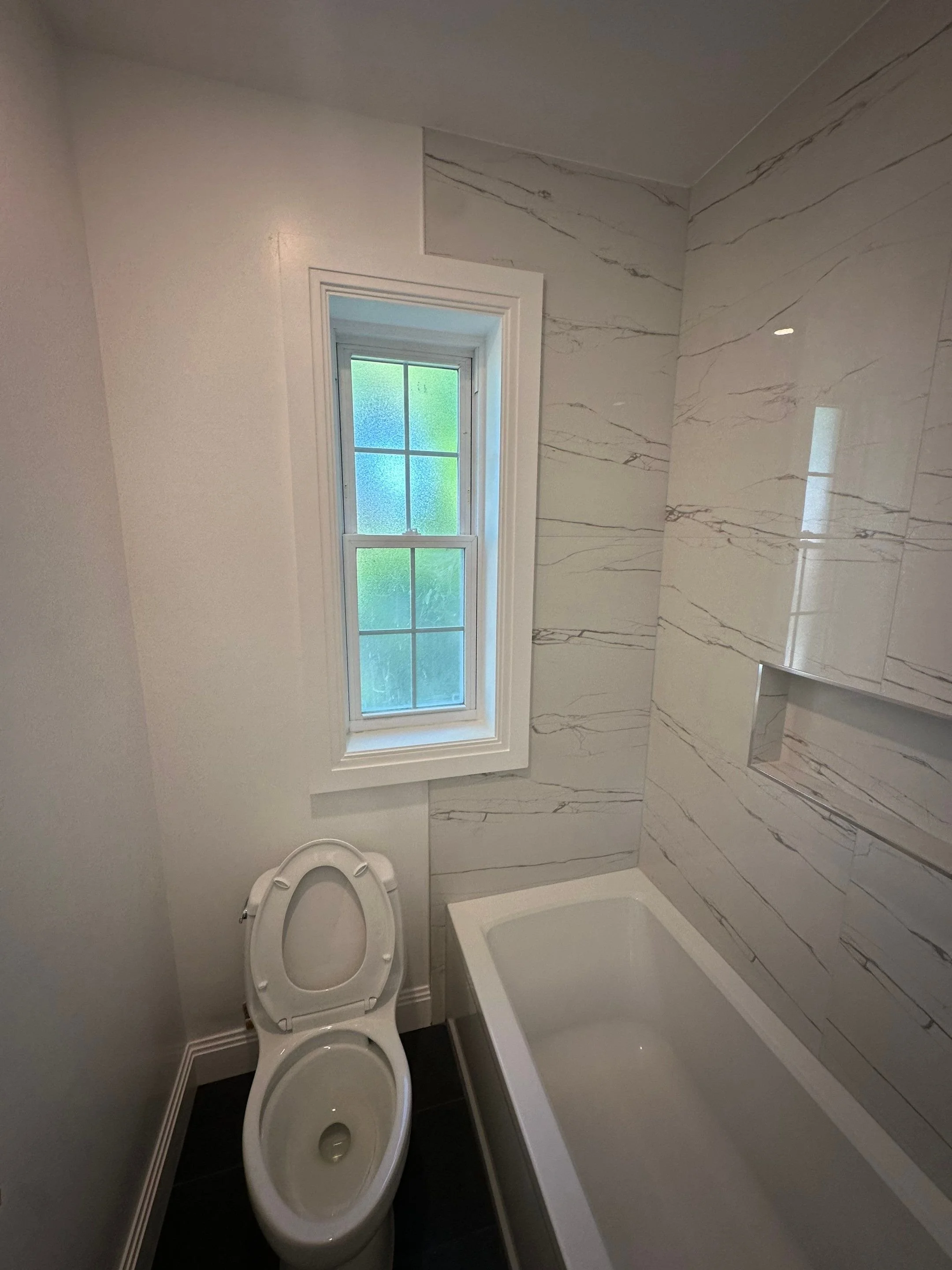 A small bathroom with a white toilet, a bathtub, and a window with frosted glass. The walls have a marble pattern.