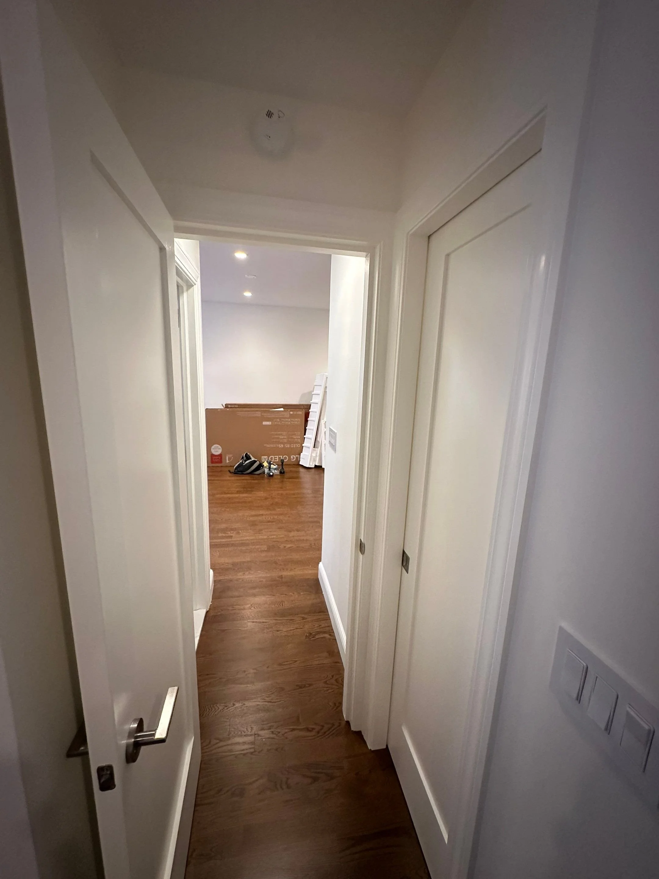 A hallway inside a house with white doors and brown wooden flooring, leading to a room with a cardboard box and some shoes.