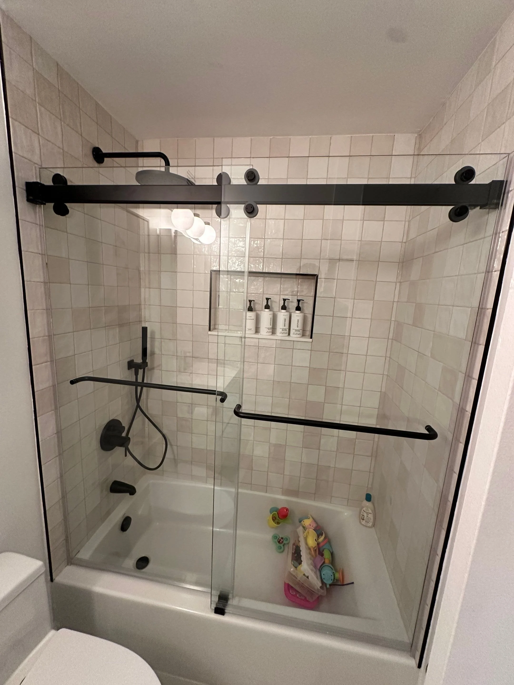 Shower with black sliding barn door, built-in niche with shampoo bottles, child's bath toys and a bottle of shampoo on the bathtub. Black fixtures, tiled walls, white bathtub.