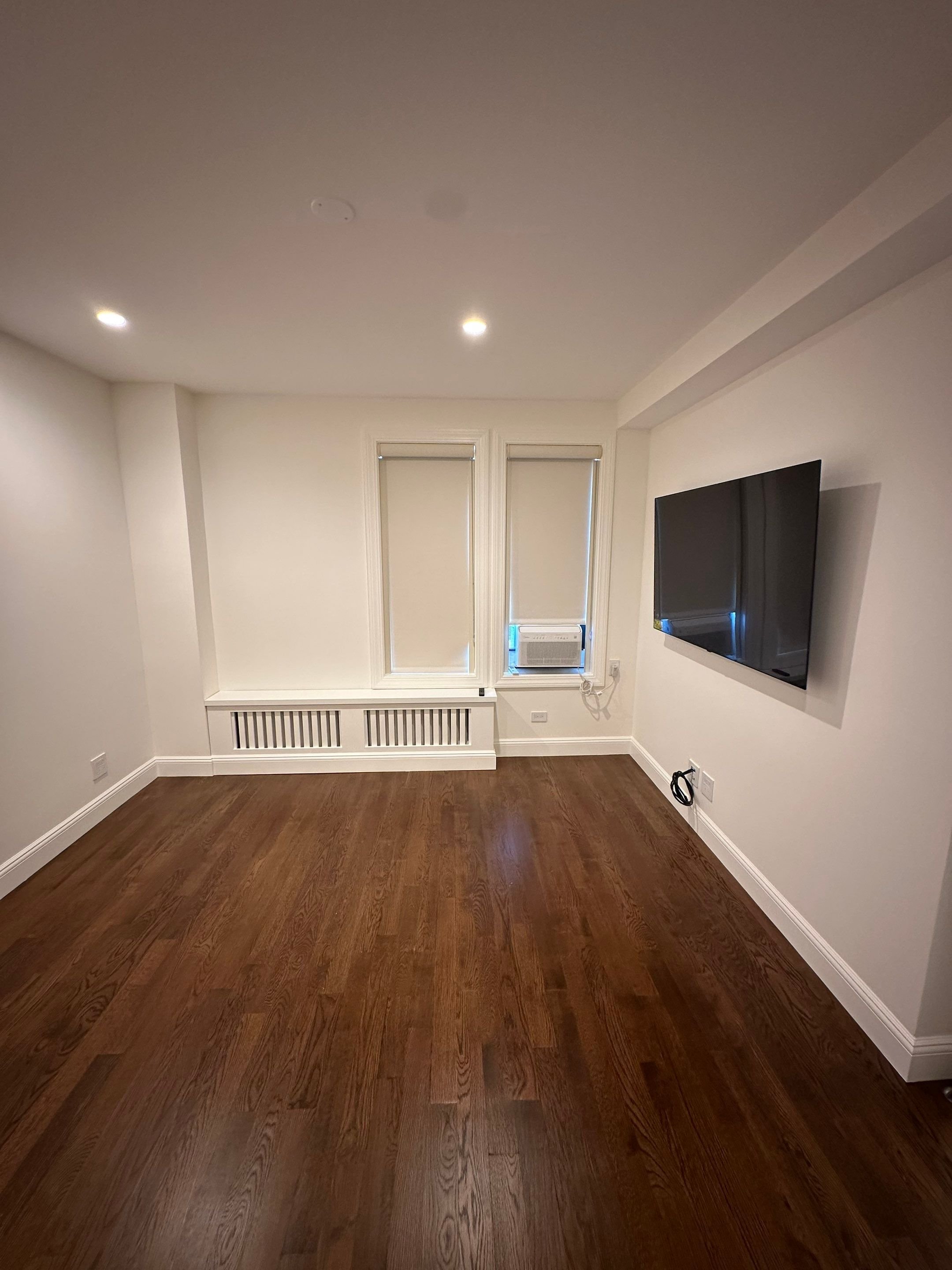 Empty living room with hardwood floors, two windows with blinds, an air conditioning unit, a mounted flat-screen TV, and recessed ceiling lights.