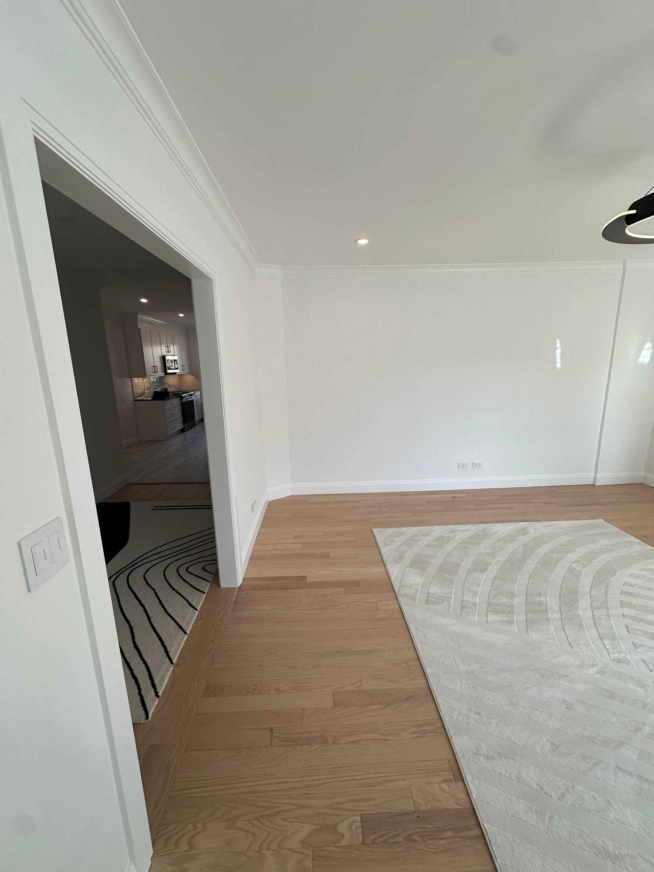 Empty room with wooden flooring, white walls, a white rug, and an open doorway leading to a kitchen.