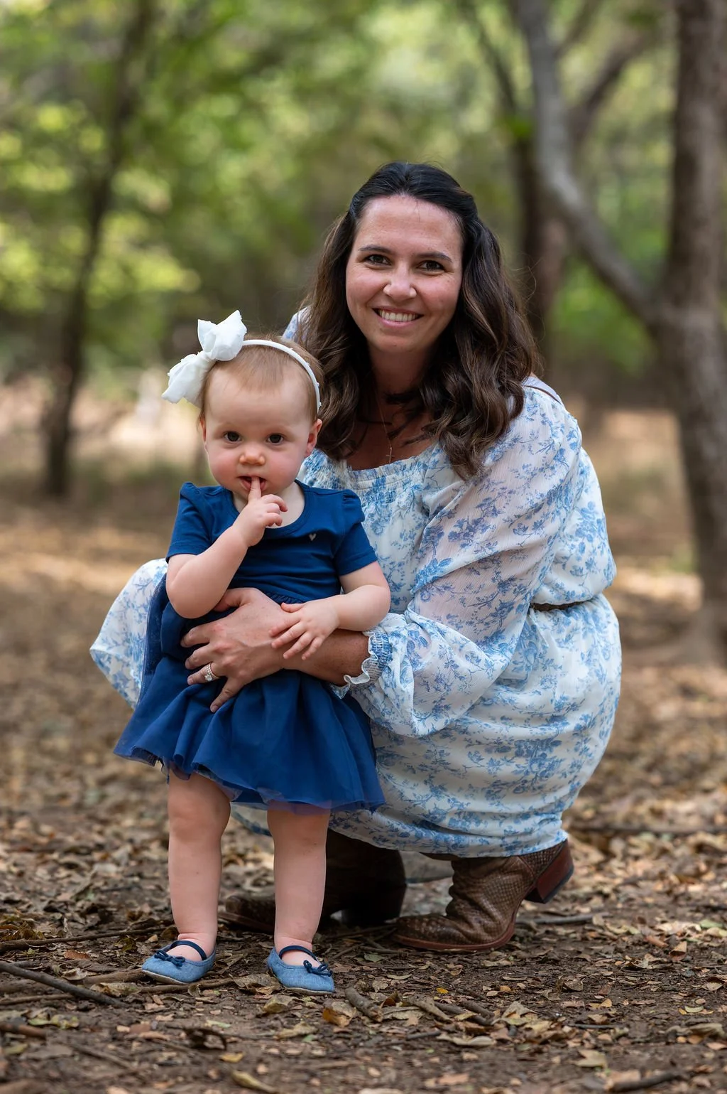 A woman with wavy dark hair and a young girl with a white bow in her hair, both wearing blue dresses, outdoors in a wooded area with green trees and leaves on the ground.