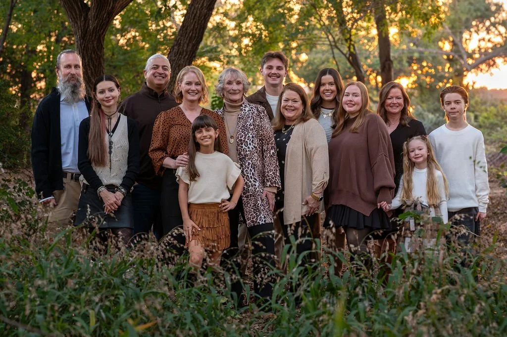 A group of fifteen people, including men, women, and children, standing outdoors in a forested area during sunset, smiling for a photo.