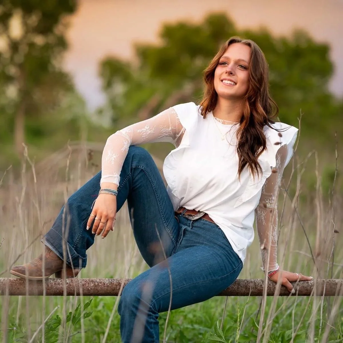 A young woman sitting on a metal railing in a rural field. She wears a white shirt with lacy sleeves, blue jeans and brown boots. 
