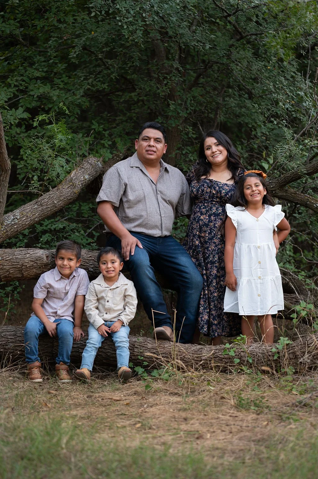 A family of five posing outdoors near a large tree with green foliage. The parents are sitting on a fallen log, with the father in a gray shirt and jeans, and the mother in a floral dress. The three children, two boys and one girl, are standing and sitting around them, dressed in casual clothes and smiling at the camera.