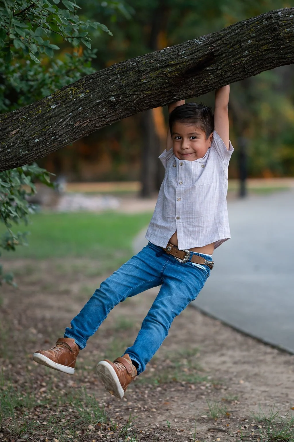 A young boy hanging from a tree branch outdoors, smiling at the camera.