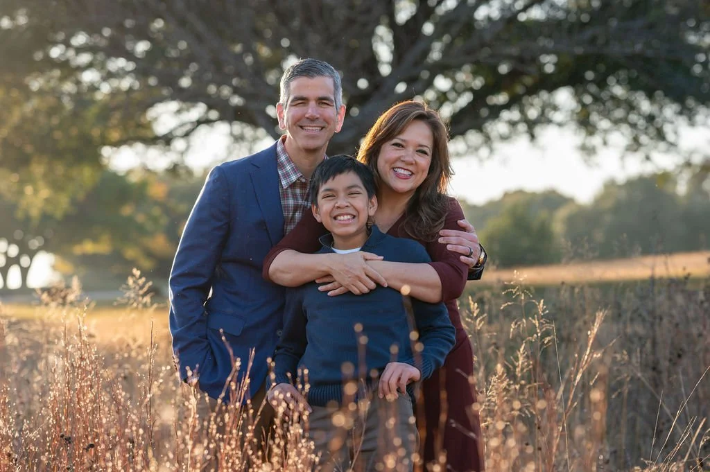 A smiling family of three standing outdoors in a field with tall grass, with trees in the background and sunlight shining through.