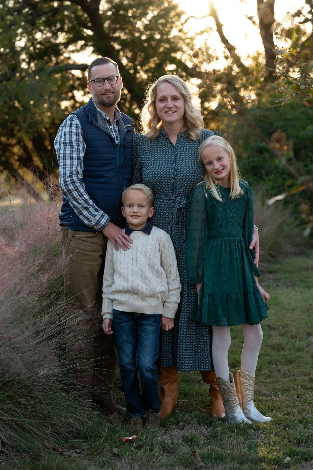 A family of five standing outdoors at sunset, with trees and greenery in the background. The father has glasses and a beard, wearing a plaid shirt and a blue vest. The mother has blonde hair, wearing a patterned dress. The children include a young boy in a cream sweater, and a girl in a green dress and white tights with cowboy boots. The family is smiling and posing for the photo.