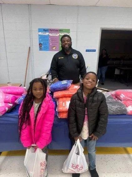 A man standing behind a table with stacks of donated clothes, with two young girls in front holding grocery bags, in an indoor setting that appears to be a donation or community center.