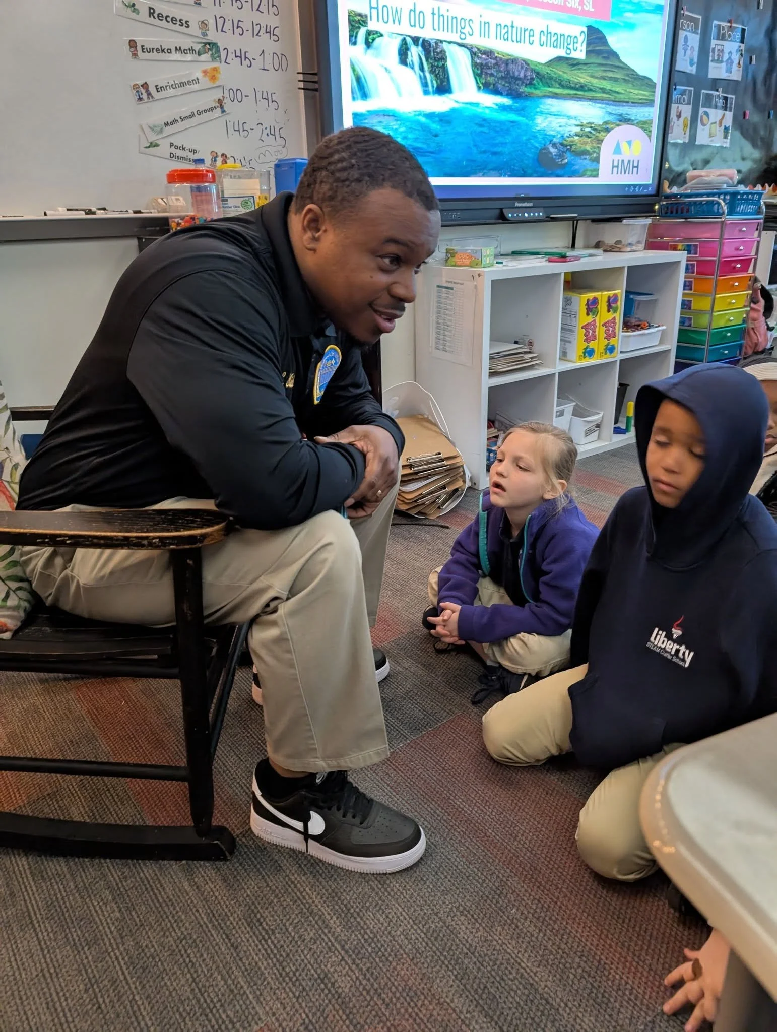 A teacher or adult engaging with two young students sitting on the classroom floor during a lesson.